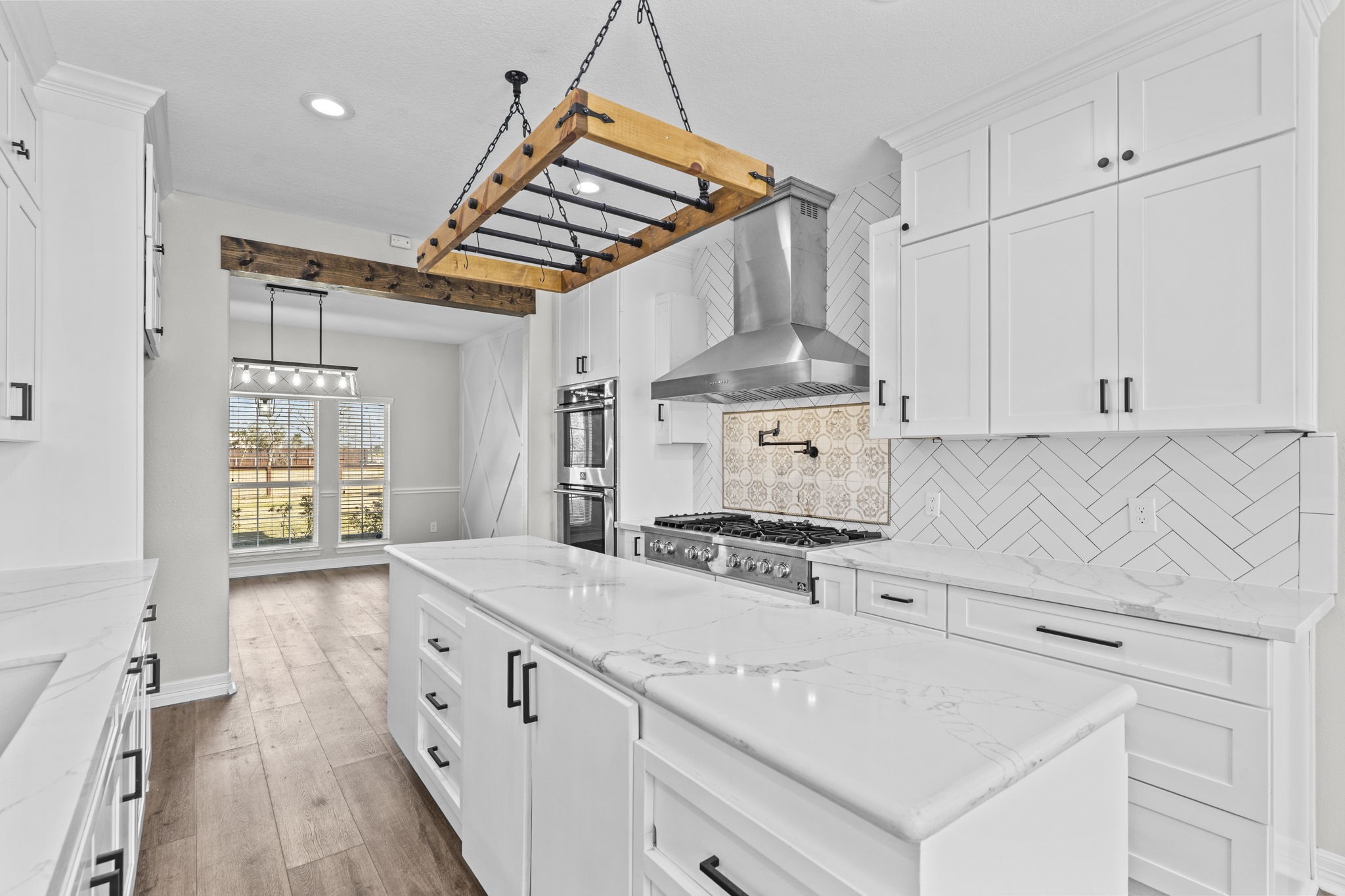 2730 Lawrence Road Kemah, TX 77565 - Photo 16 of 46 This kitchen features sleek white cabinetry, a large marble island, and modern stainless steel appliances. The herringbone backsplash and wooden beams add a touch of rustic charm. Natural light flows through the adjacent dining area, creating a bright and inviting space.