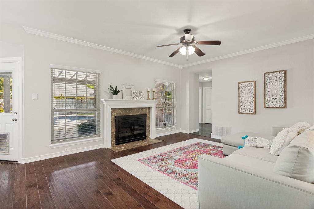 1228 Concho Drive Benbrook, TX 76126 - Photo 11 of 39 Living area featuring dark wood-finish flooring, a fireplace with a stone surround and white mantel, crown molding, a ceiling fan with light fixture, and multiple windows fitted with blinds