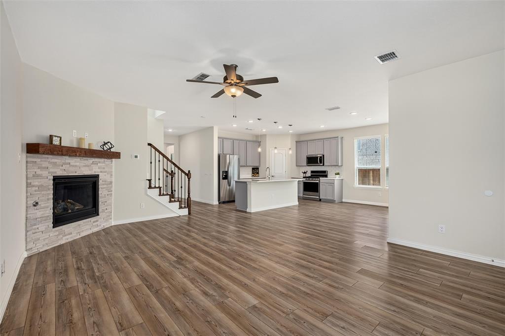 8715 Royal Star Road Rowlett, TX 75089 - Photo 13 of 39 Wood-finish flooring throughout, stacked stone fireplace with a wood mantle, ceiling fan with integrated lighting, kitchen featuring gray cabinetry and stainless steel appliances, and a staircase with wrought iron balusters