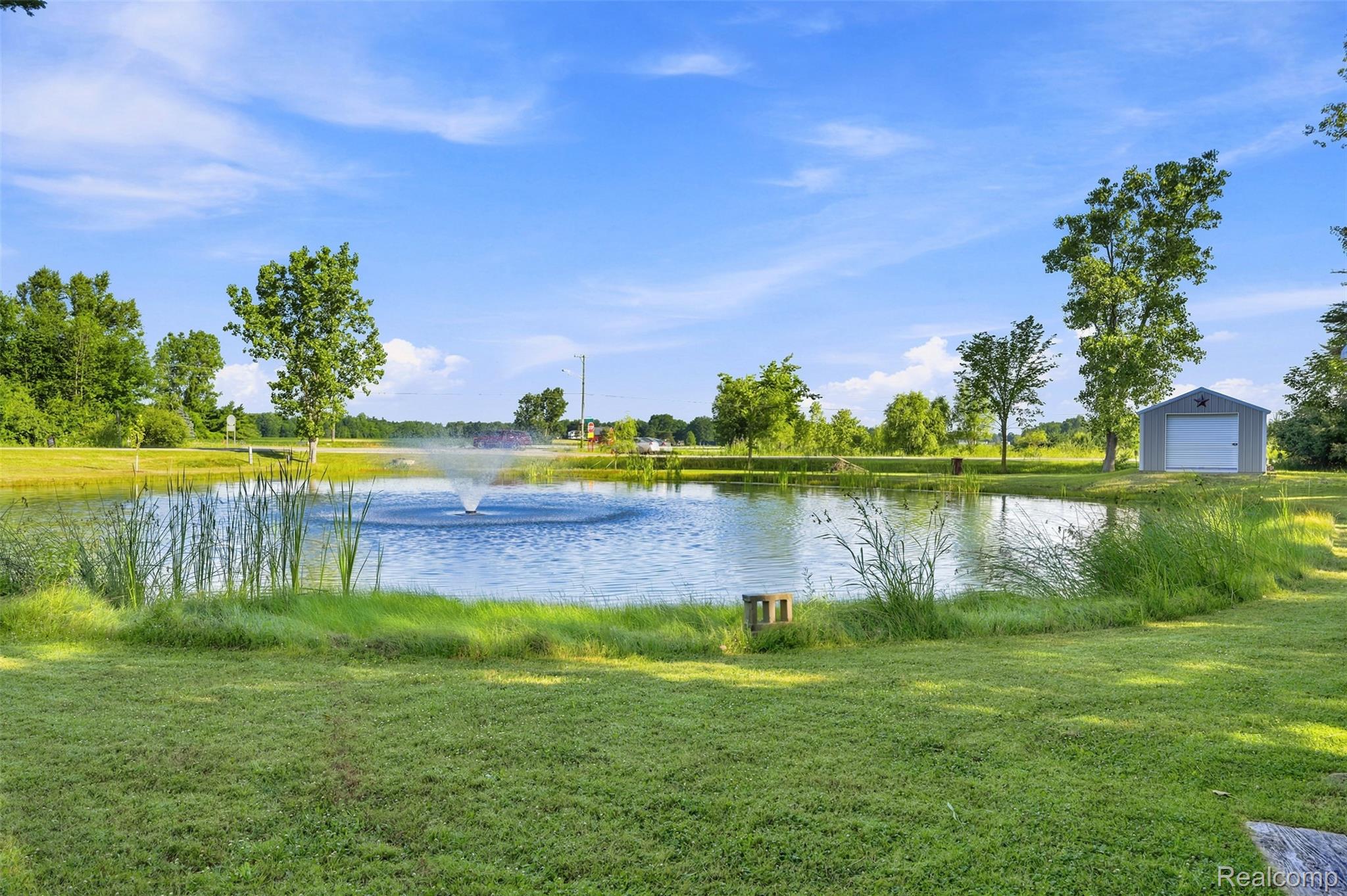 3744 Fish Lake Road Lapeer, MI 48446 - Photo 34 of 50 Expansive pond featuring a central fountain, surrounded by lush green grass, mature trees, and a gray storage shed with a white garage door