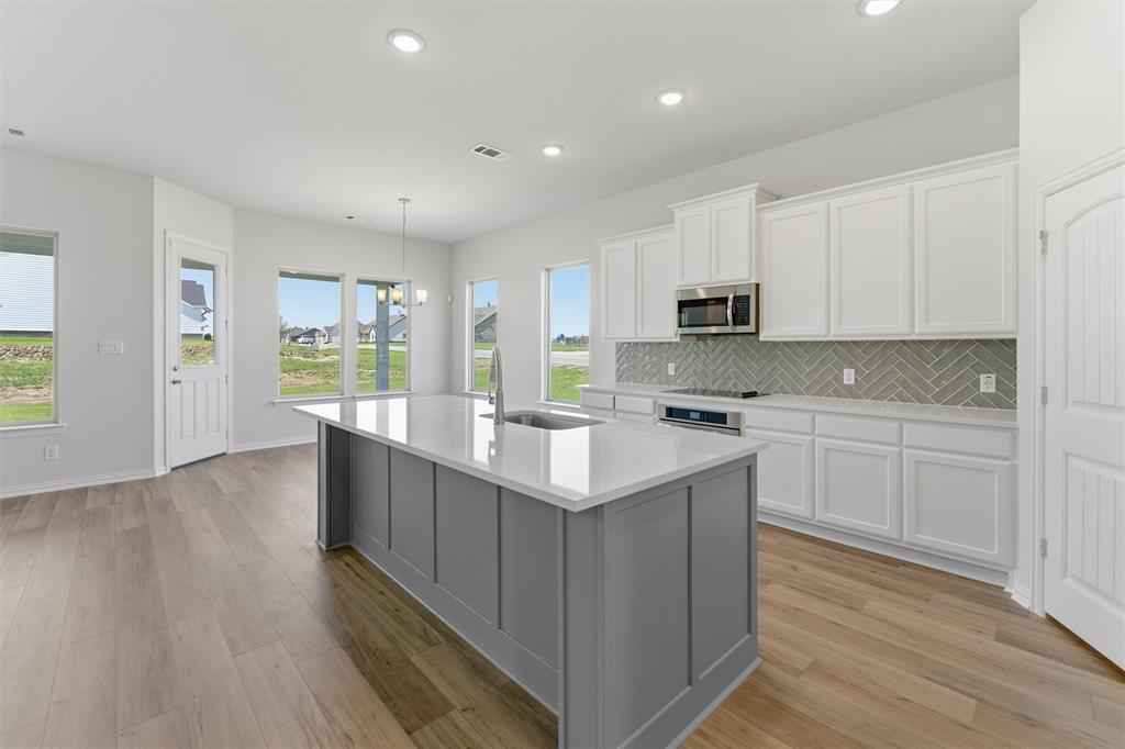 104 Snowy Owl Trail Rhome, TX 76078 - Photo 11 of 27 Open-concept kitchen featuring a large island with a sink, white cabinetry, integrated stainless steel appliances, a herringbone tile backsplash, and light wood-finish flooring