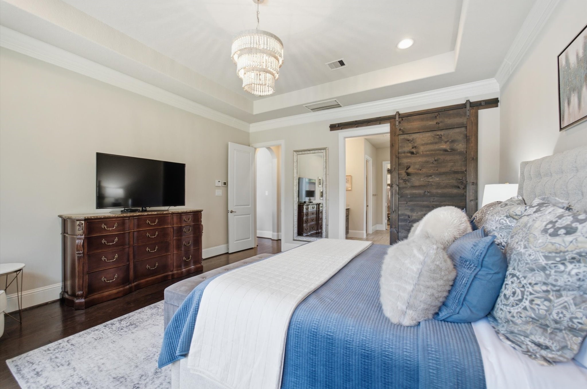 23722 Thortons Park Lane Spring, TX 77389 - Photo 12 of 48 This elegant bedroom features a tray ceiling, a stylish chandelier, and a cozy ambiance. It includes a large bed with plush pillows, a classic wooden dresser with a TV, and a rustic sliding barn door leading to an adjacent room.