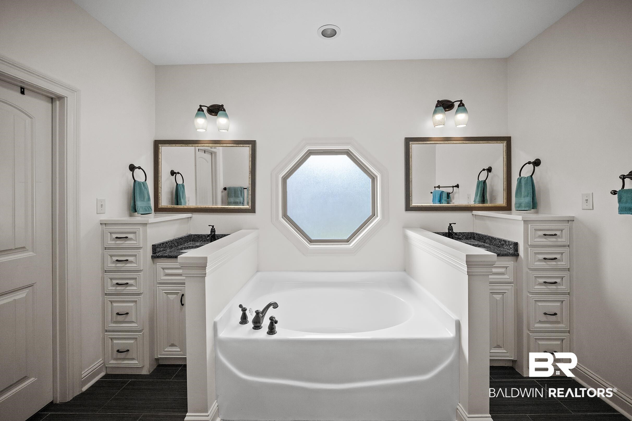 313 Pecan Ridge Boulevard Fairhope, AL 36532 - Photo 14 of 51 White soaking tub with dark bronze fixtures, flanked by dual vanities featuring black countertops and white cabinetry with drawer stacks
