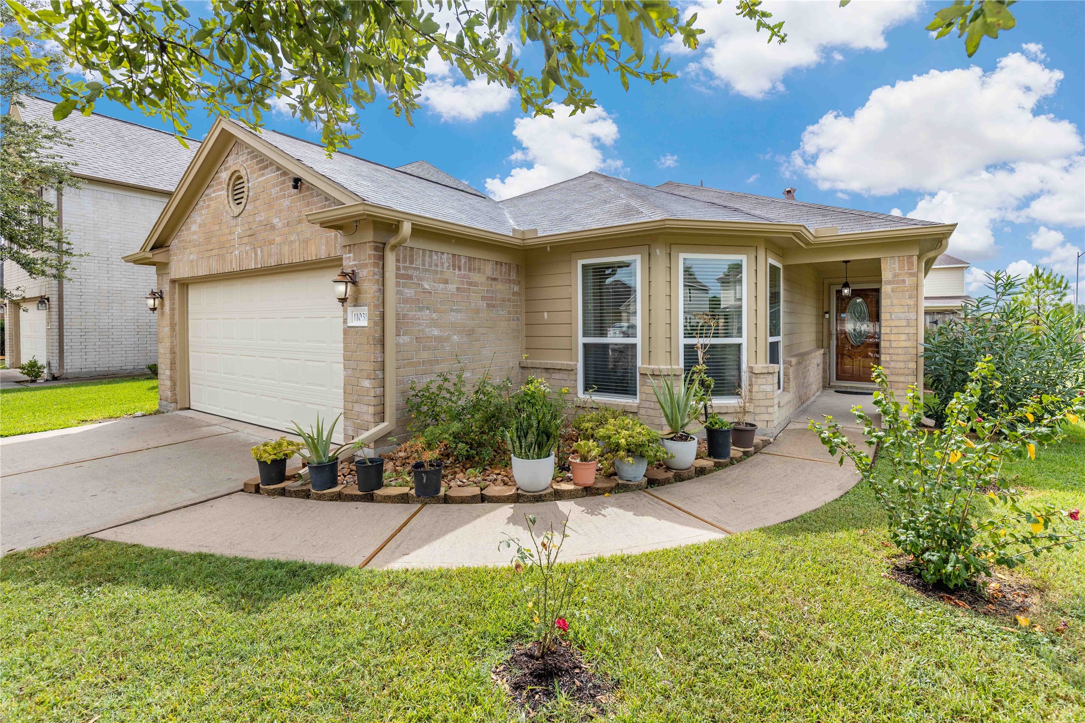 This charming single-story home features a brick exterior with a welcoming front porch. It has a two-car garage and a well-maintained front yard adorned with various potted plants. The bay window adds character to the facade, and the lush green lawn enhances curb appeal.