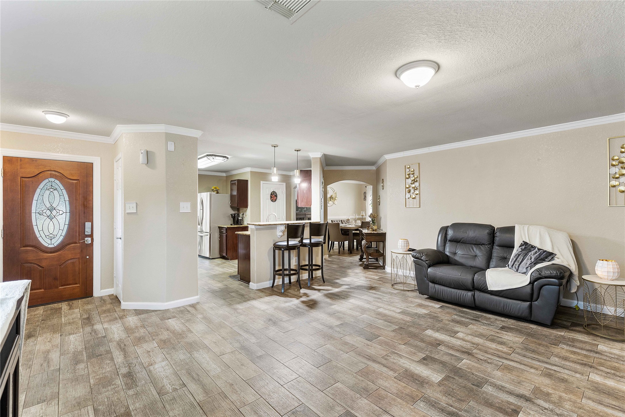 11031 Bauer Elm Street Houston, TX 77044 - Photo 12 of 33 This open-concept living area features a warm, inviting space with wood-look tile flooring, a cozy seating area, and neutral walls. The modern kitchen includes stainless steel appliances and a breakfast bar, flowing seamlessly into the dining area. The front door showcases an elegant glass design, adding character to the entryway.