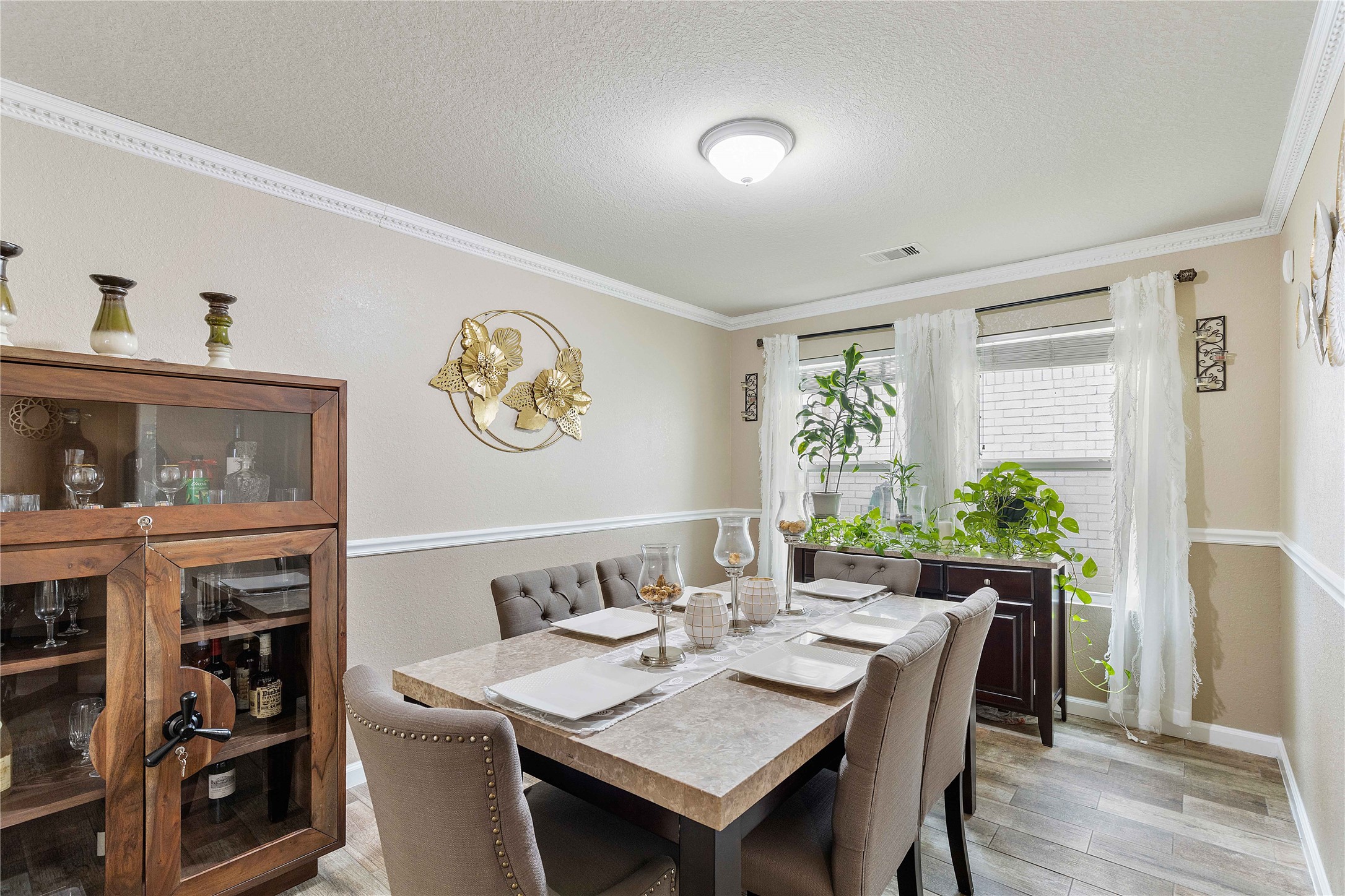 11031 Bauer Elm Street Houston, TX 77044 - Photo 15 of 33 A cozy dining room with elegant decor, featuring a marble table set for six, a wooden cabinet, and natural light from large windows with sheer curtains. The space is accented by wall art and indoor plants, creating a warm, inviting atmosphere.