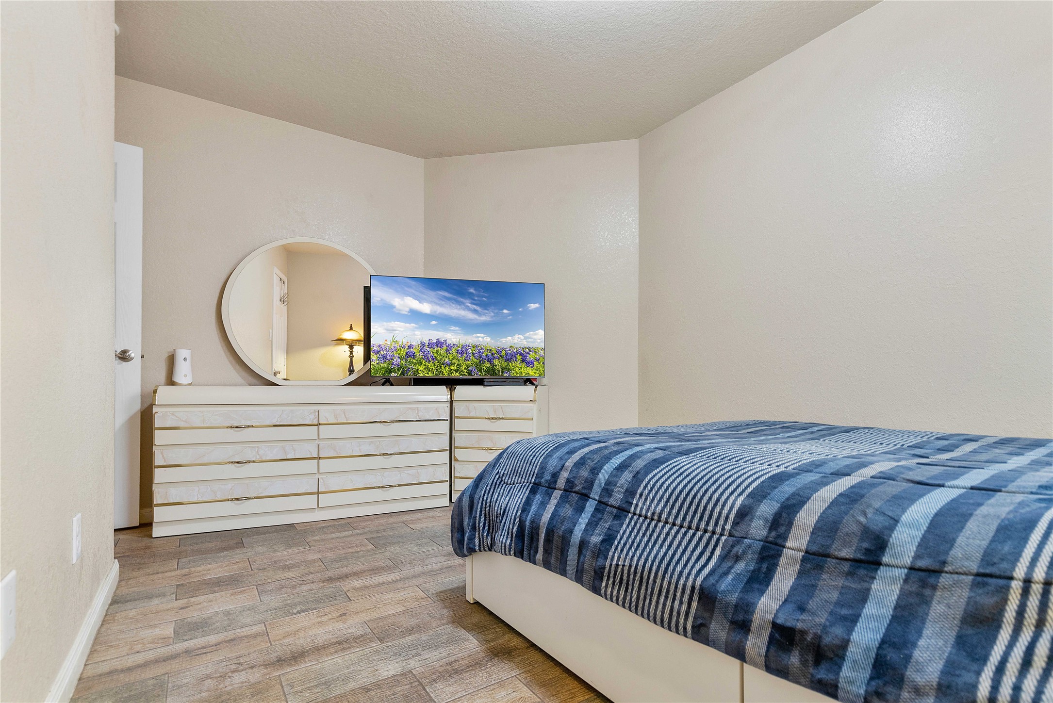 11031 Bauer Elm Street Houston, TX 77044 - Photo 19 of 33 Cozy bedroom with neutral walls features a comfortable bed with a blue plaid comforter. A large dresser with a round mirror sits against the wall, topped with a TV. The room has wood-like flooring and a simple, clean aesthetic.