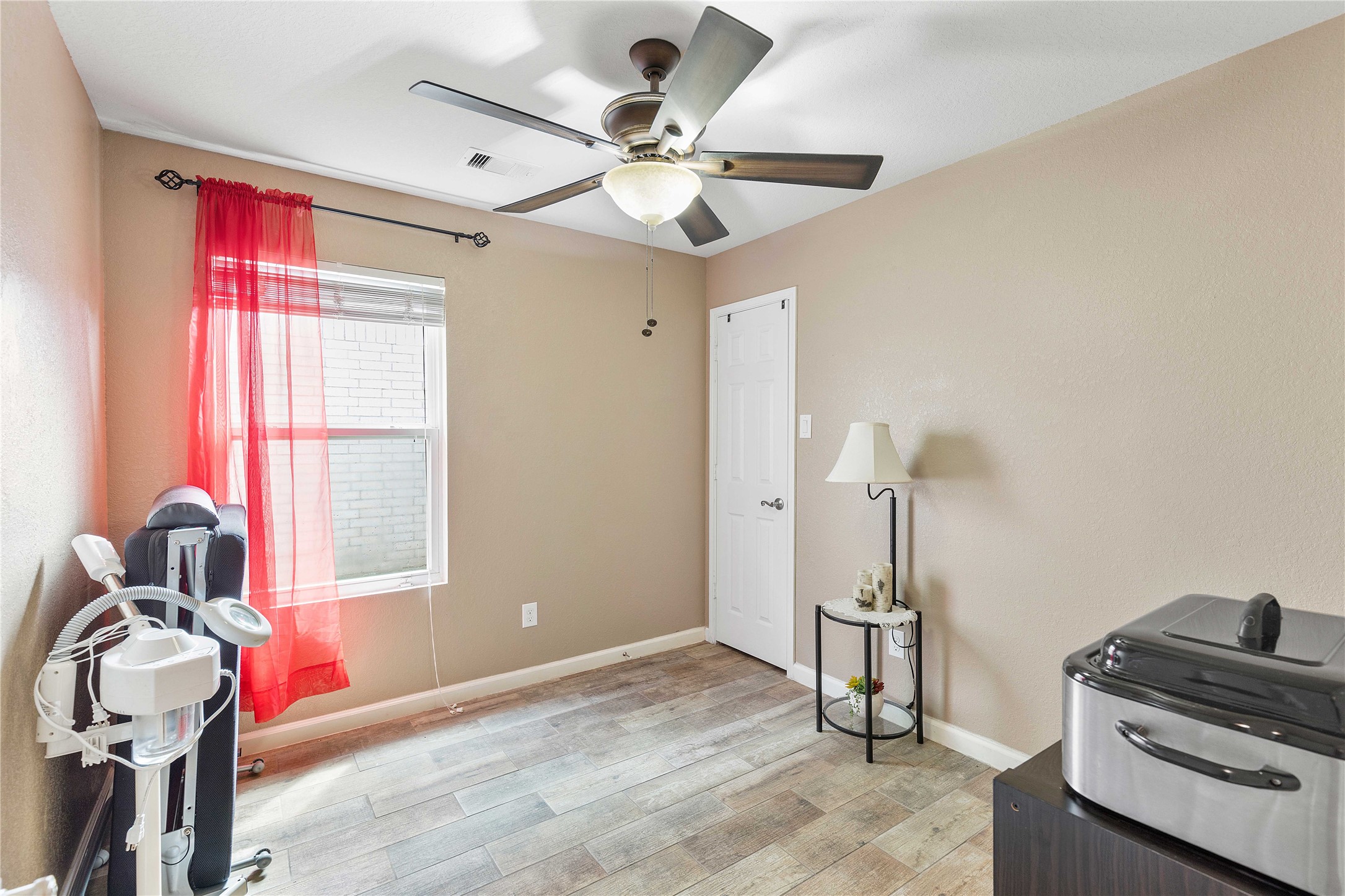 11031 Bauer Elm Street Houston, TX 77044 - Photo 20 of 33 A cozy room with neutral walls, wood-style flooring, and a ceiling fan. It features a window with red curtains, a small table with a lamp, and a closet door. Ideal for use as a small bedroom or office space.