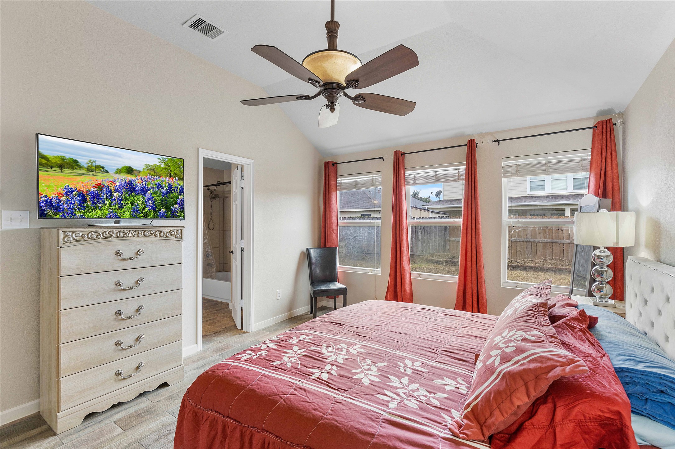 11031 Bauer Elm Street Houston, TX 77044 - Photo 25 of 33 This cozy bedroom features a vaulted ceiling with a ceiling fan, large windows with red curtains offering plenty of natural light, and a neutral color palette. It includes a plush bed, a stylish dresser, and a TV. The attached bathroom adds convenience, and the overall space exudes warmth and comfort.