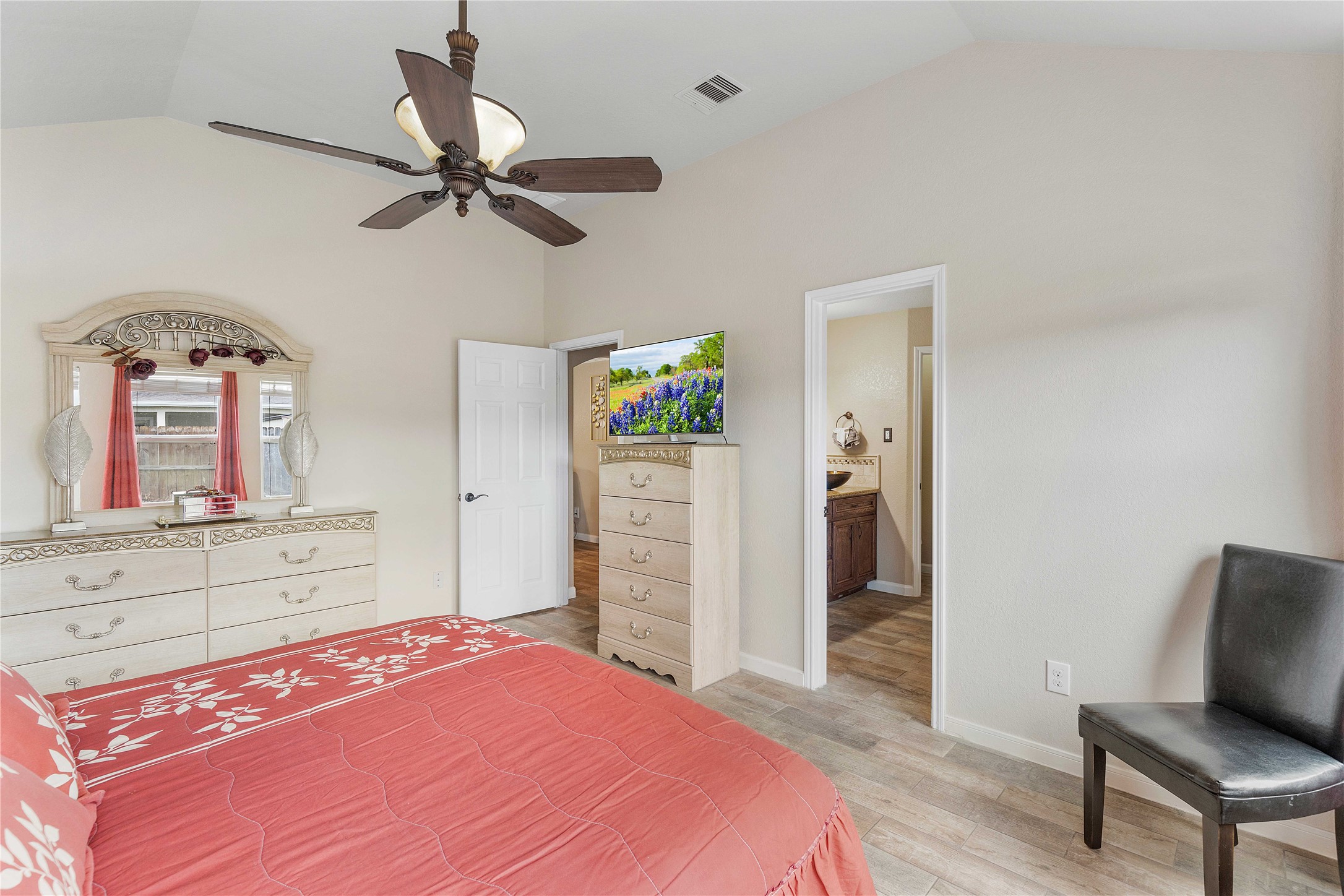 11031 Bauer Elm Street Houston, TX 77044 - Photo 26 of 33 This bedroom features a cozy atmosphere with neutral walls and wood flooring. It includes a ceiling fan, a dresser with a mirror, and a comfortable bed with a red floral bedspread. The room has an en-suite bathroom and a small TV on a chest of drawers, offering convenience and style.