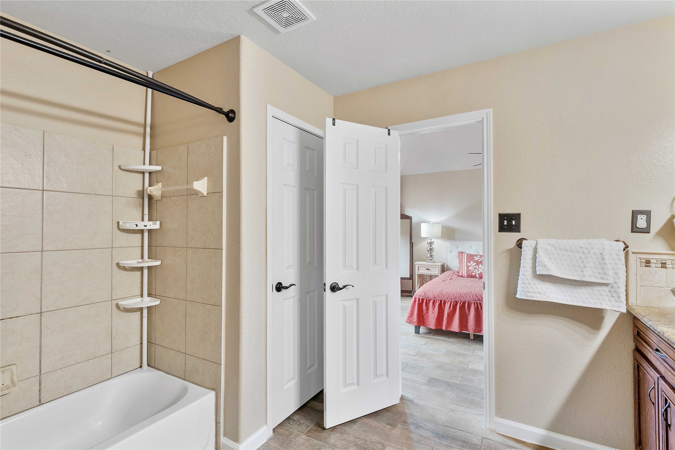 11031 Bauer Elm Street Houston, TX 77044 - Photo 28 of 33 This photo shows a bathroom with tiled walls and a shower-tub combo. There's a towel rack and a glimpse into a bedroom featuring a bed with a red comforter. The space is clean and well-lit, offering a cozy, connected layout.