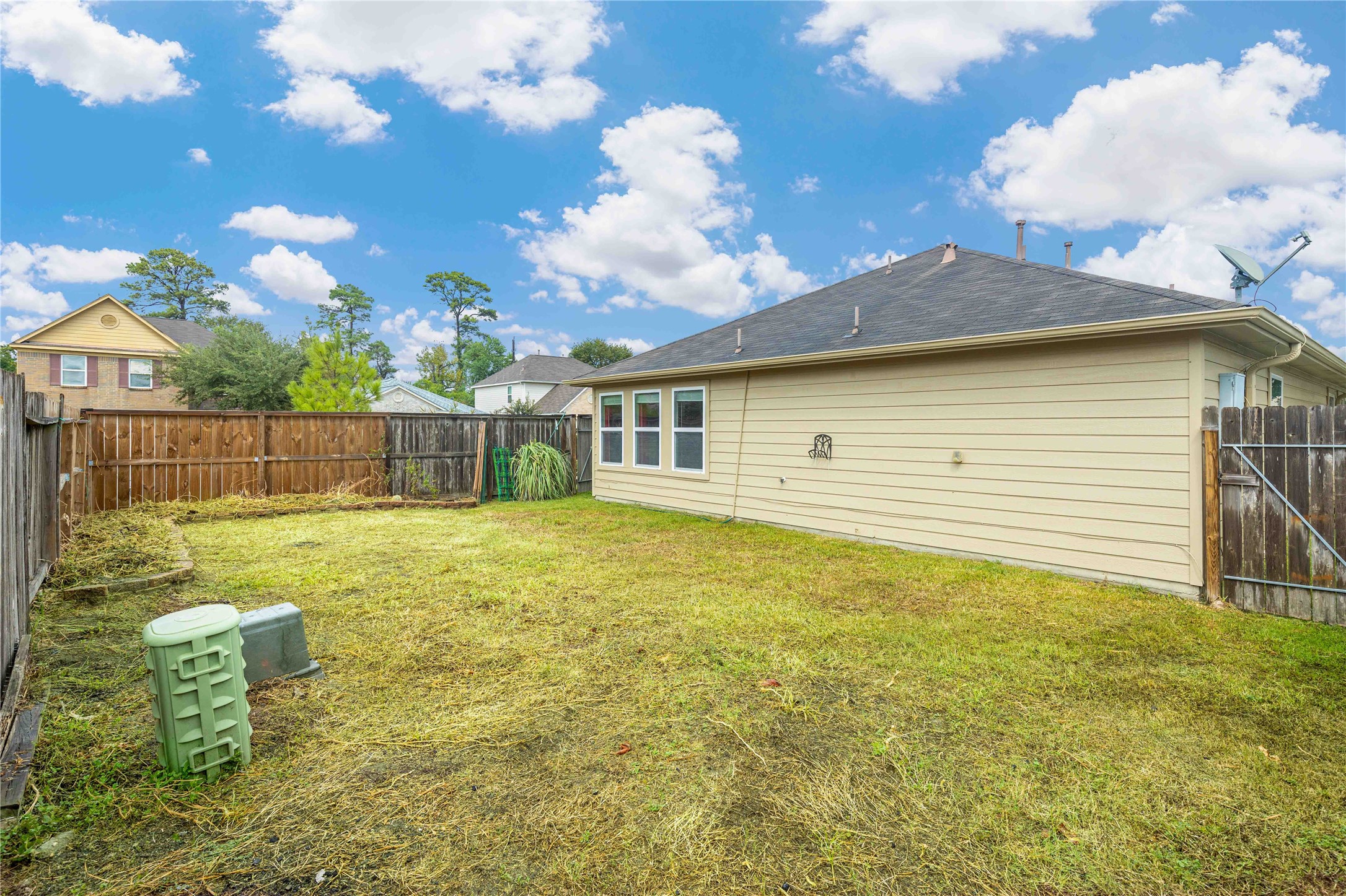 11031 Bauer Elm Street Houston, TX 77044 - Photo 30 of 33 This photo shows a fenced backyard with a small lawn area, adjacent to a single-story house. The yard has a wooden fence, some utility boxes, and a clear blue sky above, offering a private outdoor space.