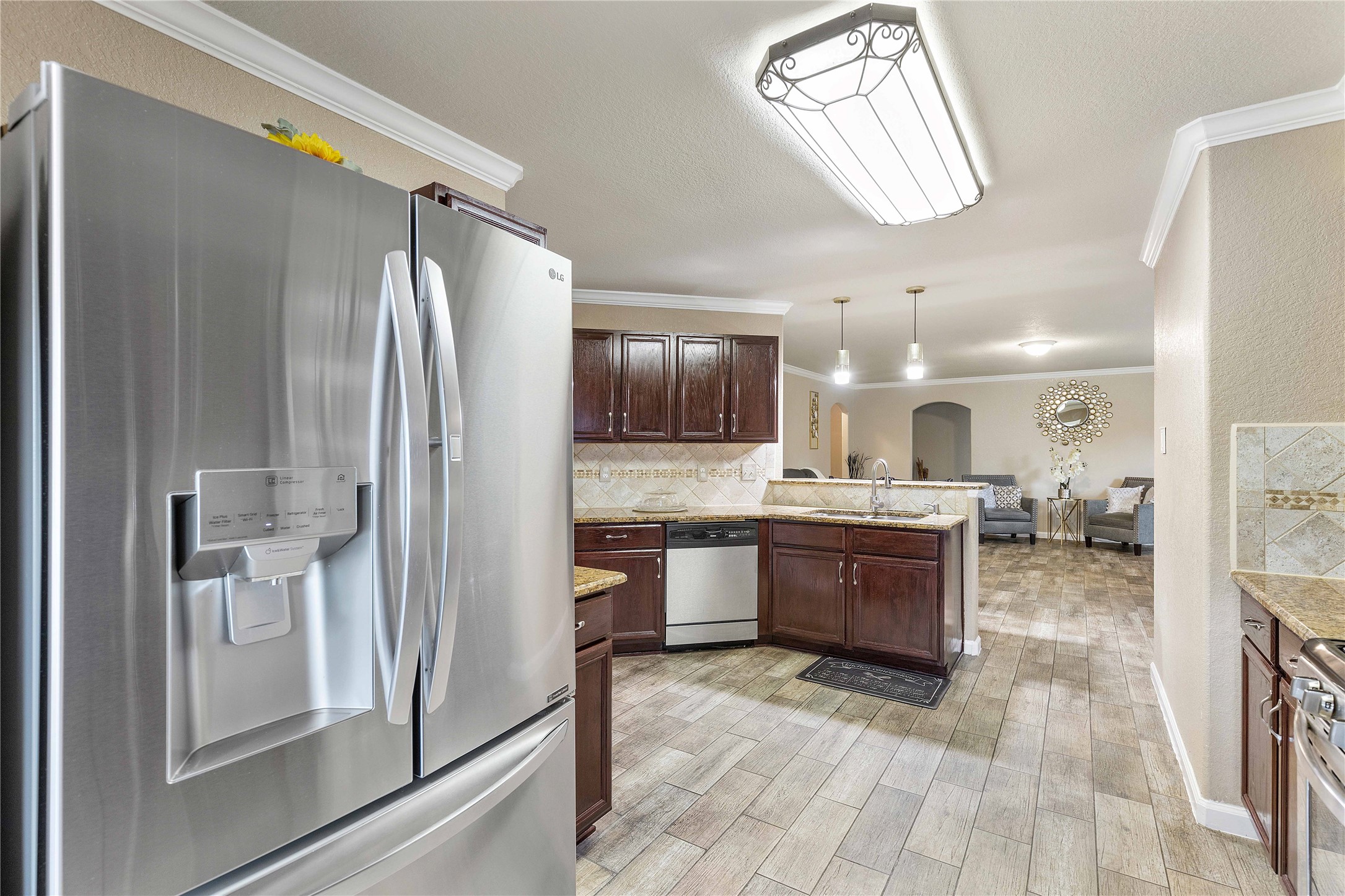 11031 Bauer Elm Street Houston, TX 77044 - Photo 9 of 33 This photo showcases a modern kitchen with stainless steel appliances, including a double-door refrigerator and dishwasher. The kitchen features dark wood cabinets, granite countertops, and a tile backsplash. It opens up to a bright, spacious living area with tiled flooring, stylish lighting fixtures, and elegant decor.