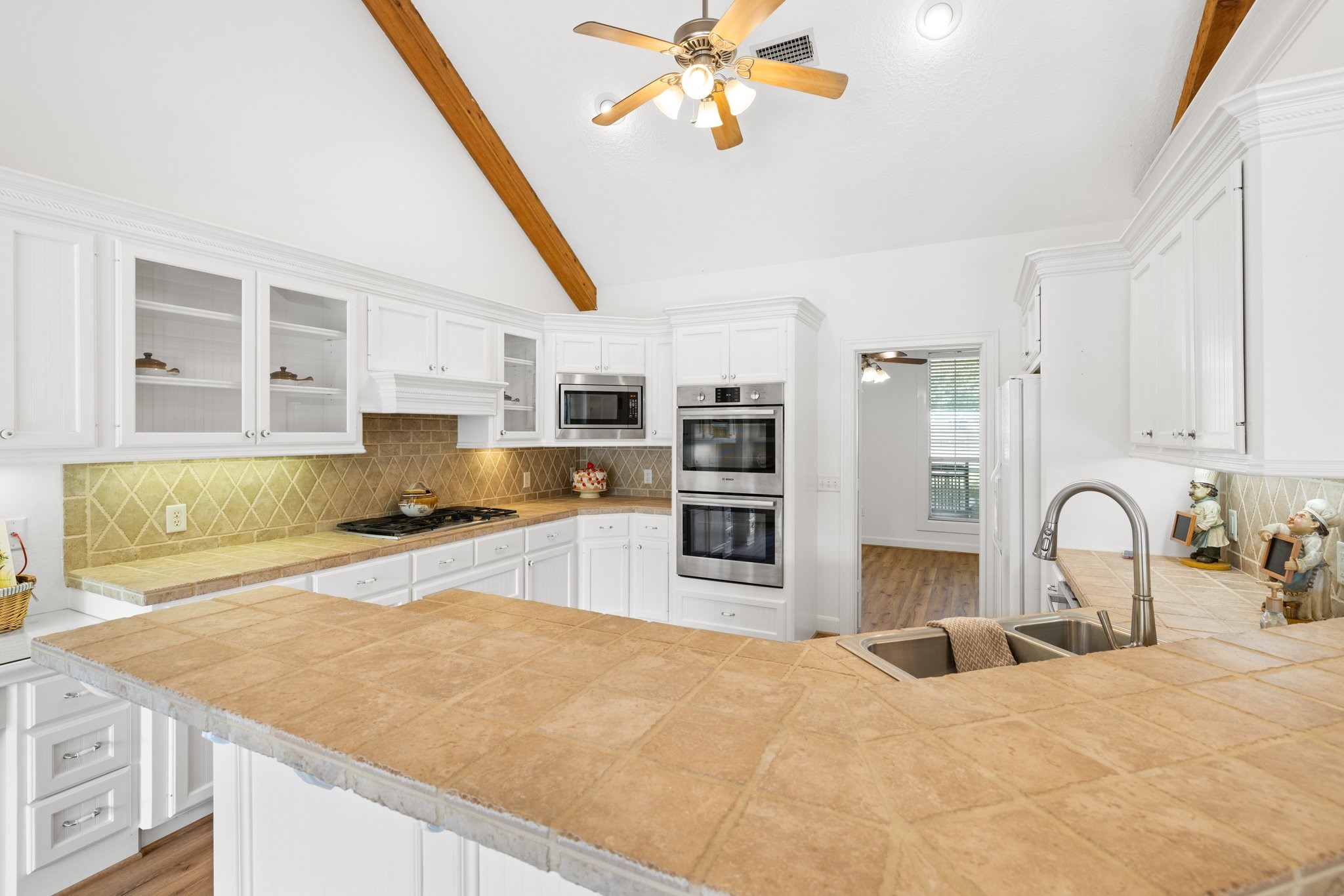 34310 High Point Drive Magnolia, TX 77355 - Photo 10 of 48 This kitchen features high vaulted ceilings with exposed wooden beams, white cabinetry, and a central island with a breakfast bar. It includes stainless steel appliances, a double oven, and a modern sink, all complemented by warm tile countertops and backsplash.