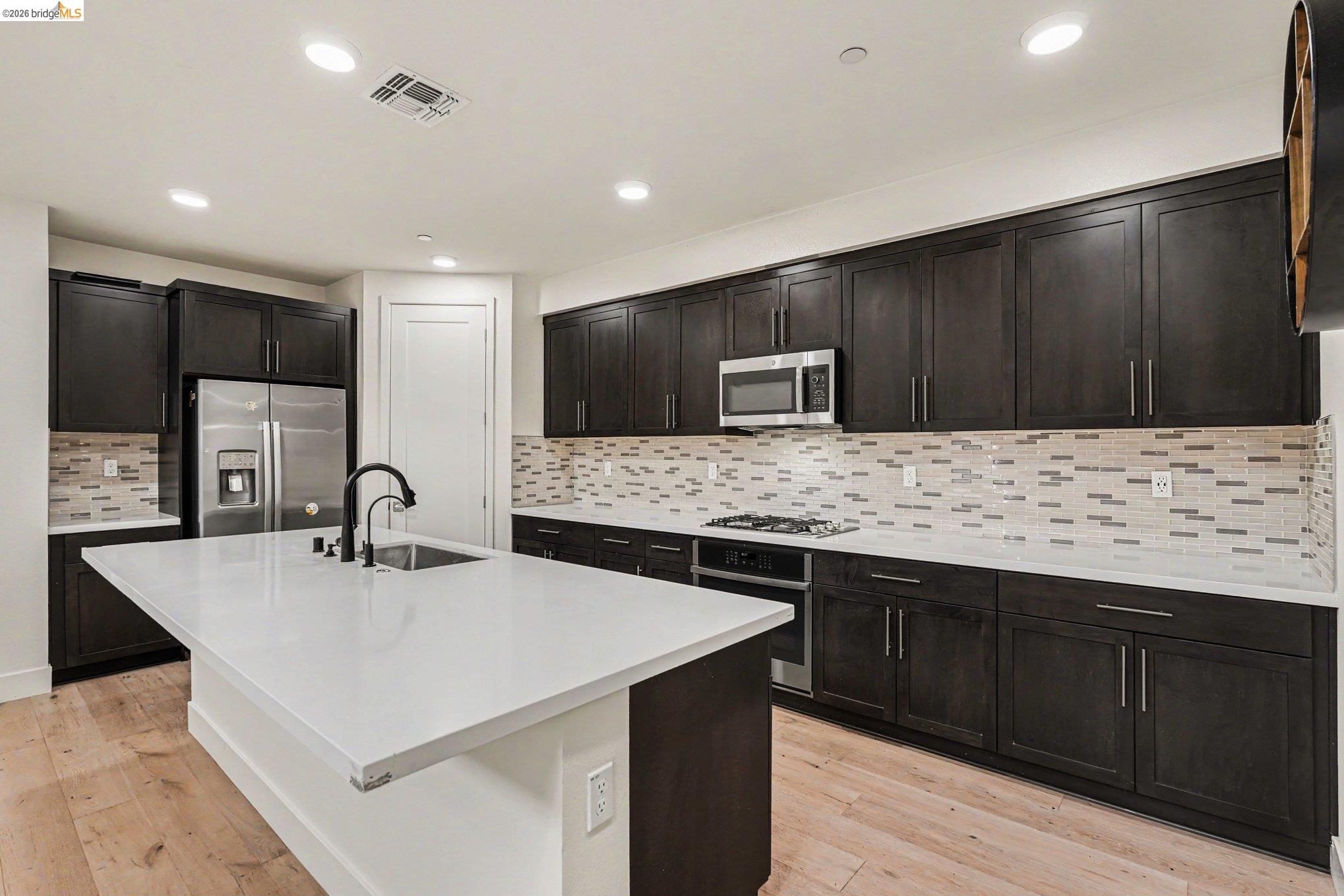 18210 Argento Loop San Ramon, CA 94583 - Photo 13 of 43 Spacious kitchen featuring an expansive island with an undermount sink and matte black faucet, dark wood-finish cabinetry, light-toned countertops, a mosaic tile backsplash, and stainless steel appliances including a refrigerator and microwave