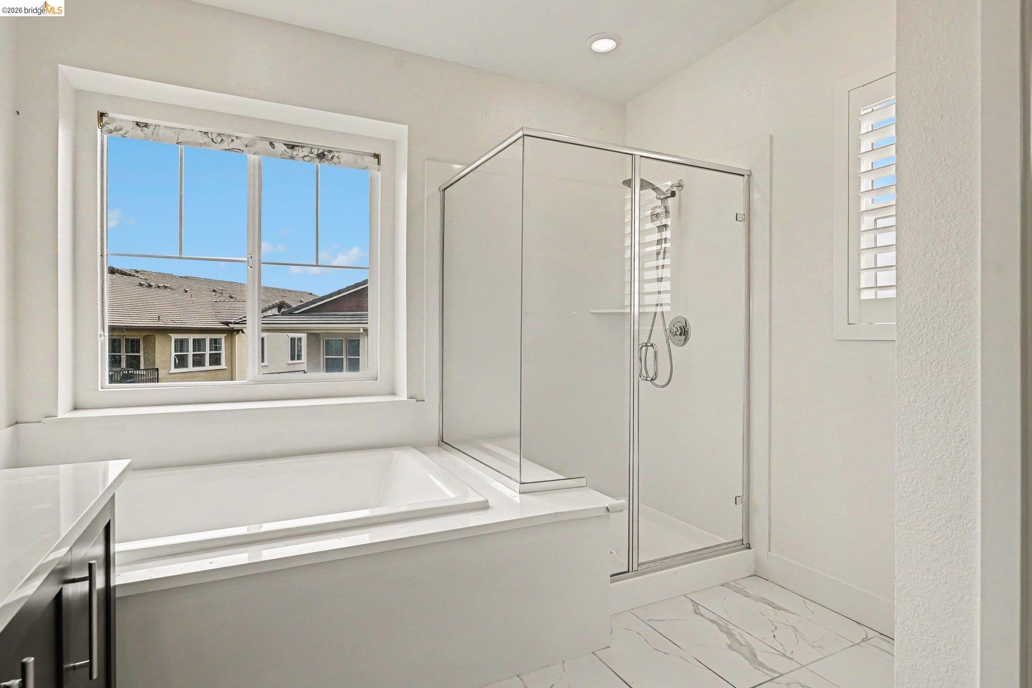 18210 Argento Loop San Ramon, CA 94583 - Photo 25 of 43 Spacious bathroom featuring a built-in soaking tub, a glass-enclosed shower, a large window, a second window with plantation shutters, and large format marble-look floor tiles