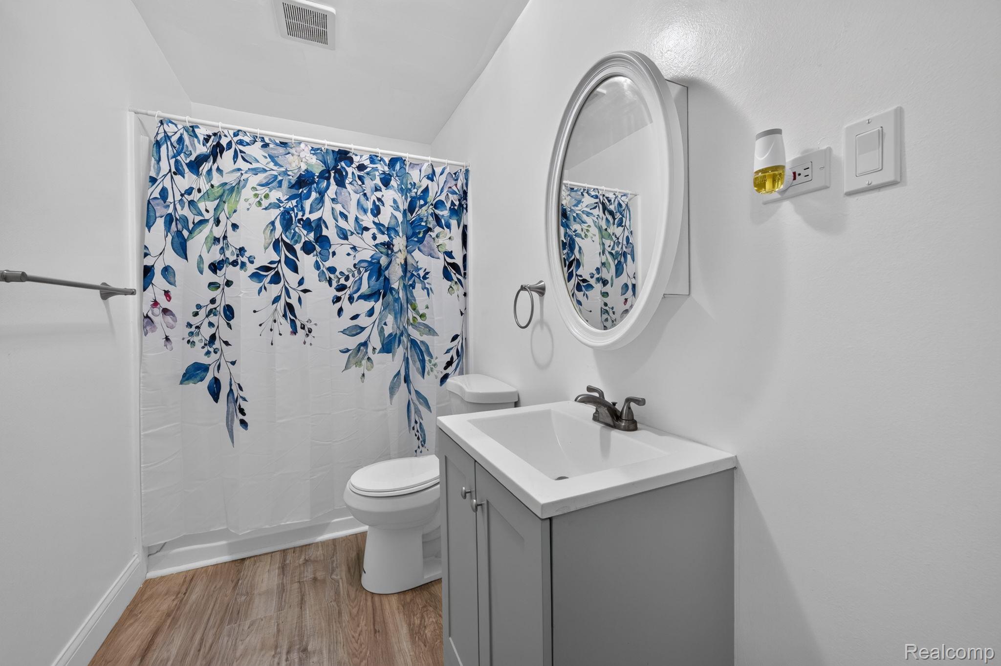 9288 Hilda Lane Flushing, MI 48433 - Photo 21 of 50 Bathroom featuring a built-in shower, wood-finish flooring, a white vanity with a rectangular sink, a wall-mounted oval mirror, and a white toilet