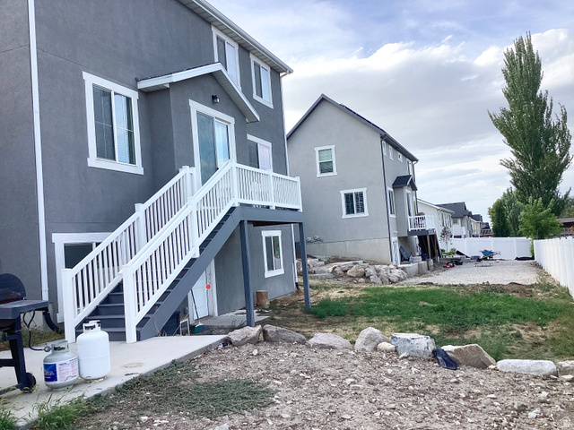 Gray stucco exterior with white trim, upper-level balcony, white railing system, lower-level concrete patio, and gravel landscaping