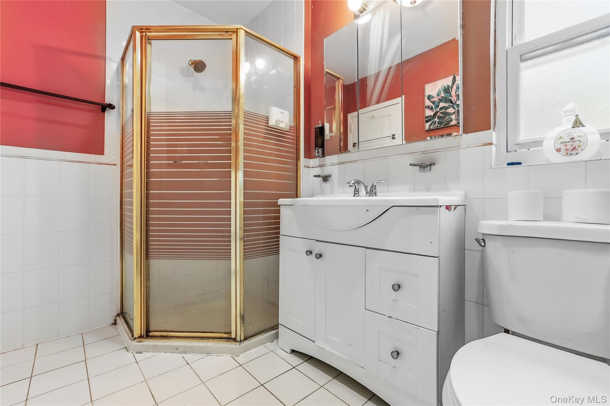 83-39 252nd Street Queens, NY 11426 - Photo 16 of 26 Bathroom featuring an enclosed corner shower with a gold-finish frame, a white vanity with integrated sink, a mirrored medicine cabinet, a window, and white square floor tiling