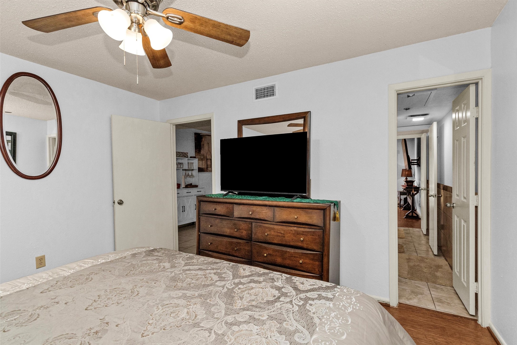 12900 Walden Road, Unit D 411 Montgomery, TX 77356 - Photo 15 of 18 This inviting bedroom features a ceiling fan, wall-mounted mirror, and dresser with a TV, seamlessly connecting to a hallway and another room, all enhanced by warm lighting for a cozy atmosphere.