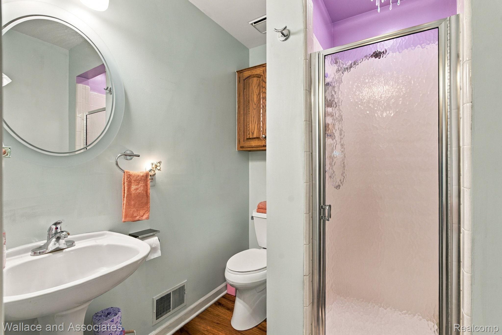 17076 27 Mile Road Ray, MI 48096 - Photo 36 of 63 Pedestal sink with chrome fixtures, round wall mirror, wood-finish flooring, enclosed shower with frosted glass, and built-in wood cabinetry
