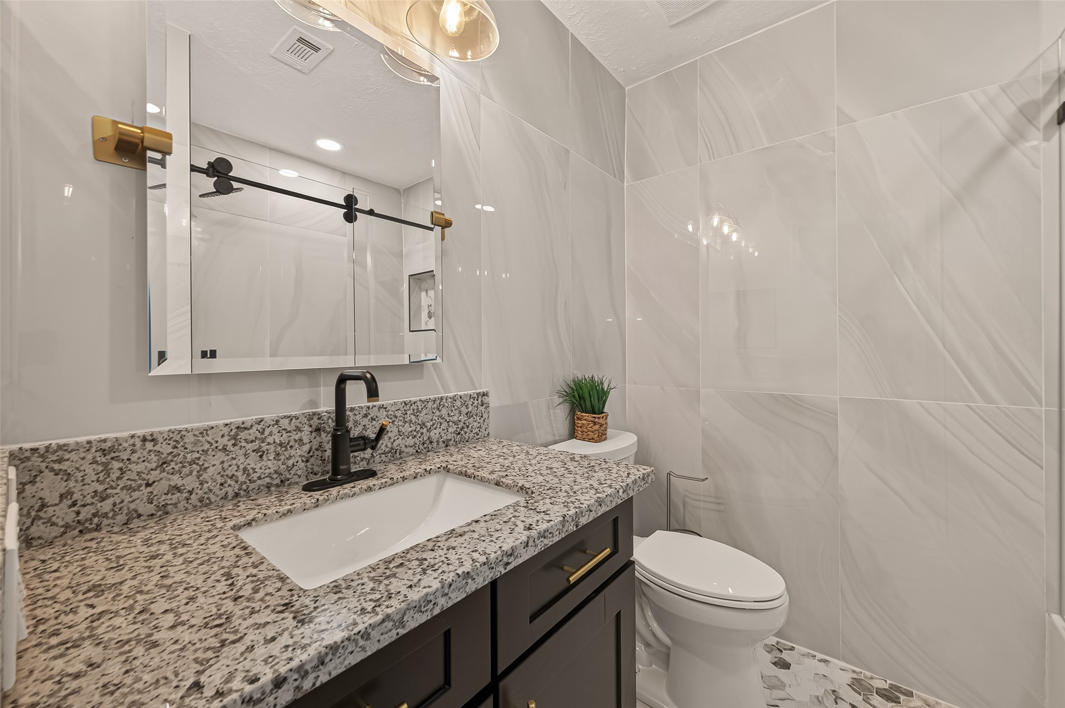 16619 Tibet Road Friendswood, TX 77546 - Photo 21 of 29 This modern bathroom features a sleek design with large marble-style wall tiles, a granite countertop, and a black faucet. The vanity has ample storage with dark cabinets and gold hardware. A stylish mirror and elegant lighting add a touch of sophistication.