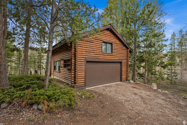 4725 Pine Ridge Road Evanston, UT 82930 - Photo 62 of 82 Log cabin exterior featuring a brown garage door, two windows with dark trim, and surrounding mature evergreen trees