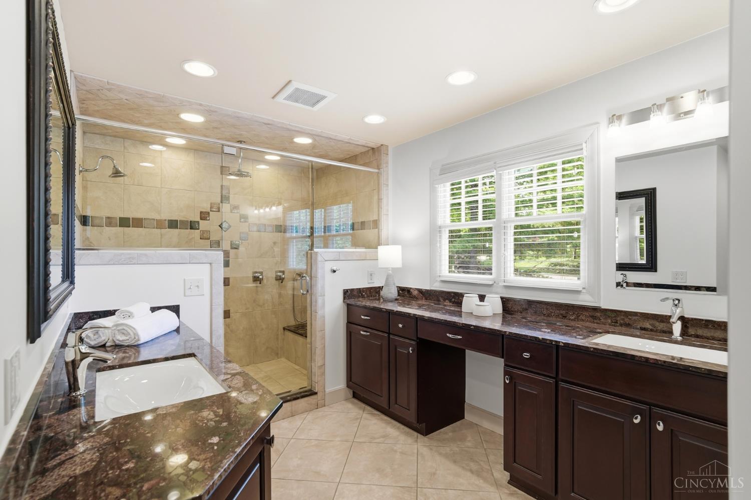 2780 Walsh Road Cincinnati, OH 45208 - Photo 36 of 77 The ensuite bath is thoughtfully designed with both comfort and functionality in mind, featuring dual vanities with rich cabinetry and expansive granite countertops that provide ample space for everyday routines. Warm, sophisticated finishes are complemented by abundant natural light, creating a bright yet inviting atmosphere. The oversized walk-in shower serves as a true focal point, showcasing full tile surround, decorative inlay accents, multiple shower heads, and a built-in bench for added convenience. A private water closet enhances the layout, offering both privacy and practicality, while generous storage throughout completes this well-appointed primary bath.