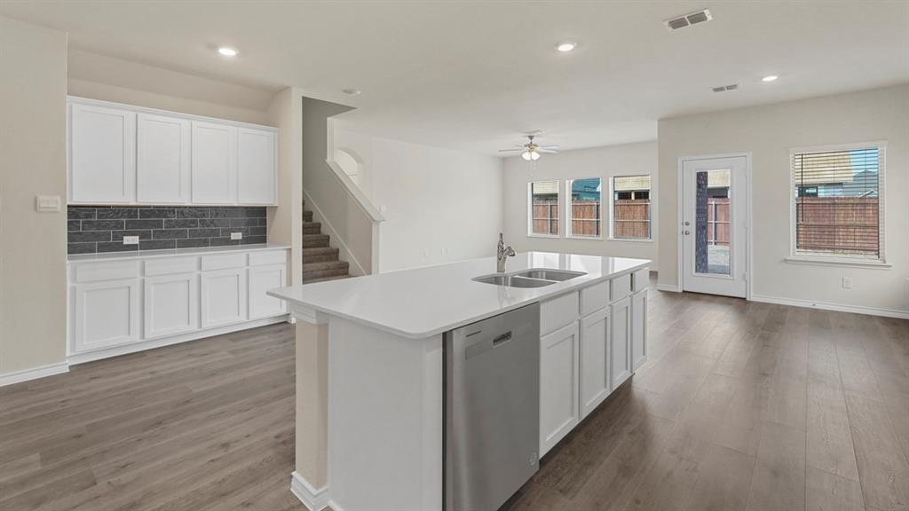447 Epiphany Lane Lavon, TX 75166 - Photo 2 of 31 Open-concept living area featuring wood-finish flooring, a kitchen island with dual sinks and dishwasher, white cabinetry with dark subway tile backsplash, and multiple windows for natural light