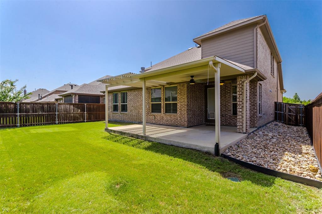 15713 Canyon Ridge Prosper, TX 75078 - Photo 36 of 38 Expansive backyard featuring a covered patio with a ceiling fan, a lush green lawn, a wooden privacy fence, and a rock-filled drainage area along the side of the home
