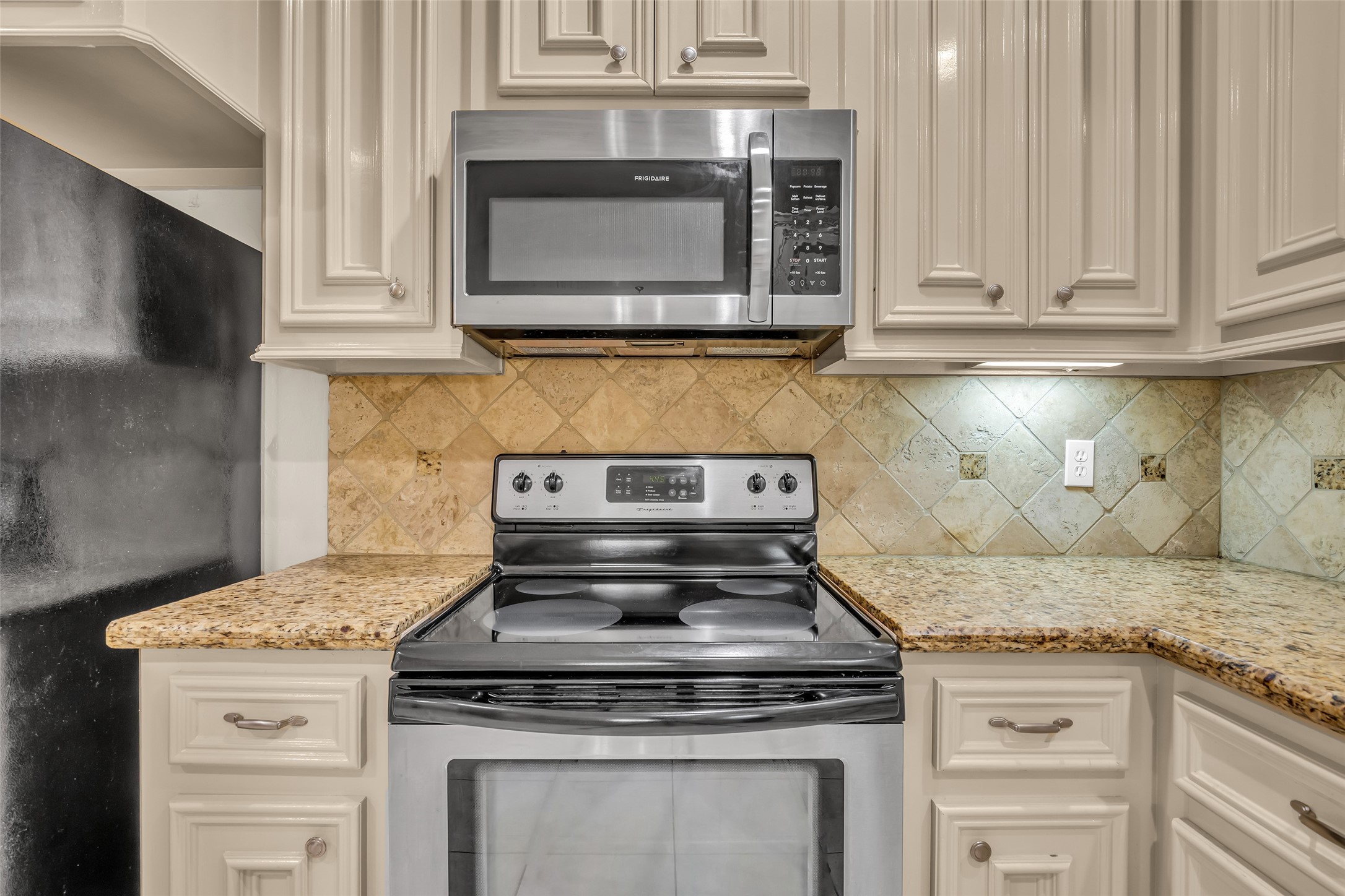 1898 Longmire Road, Unit 5 Conroe, TX 77304 - Photo 22 of 48 This photo showcases a modern kitchen corner featuring stainless steel appliances, including a microwave and oven. The space has elegant granite countertops, cream-colored cabinetry, and a tile backsplash with subtle decorative accents, offering a blend of functionality and style.