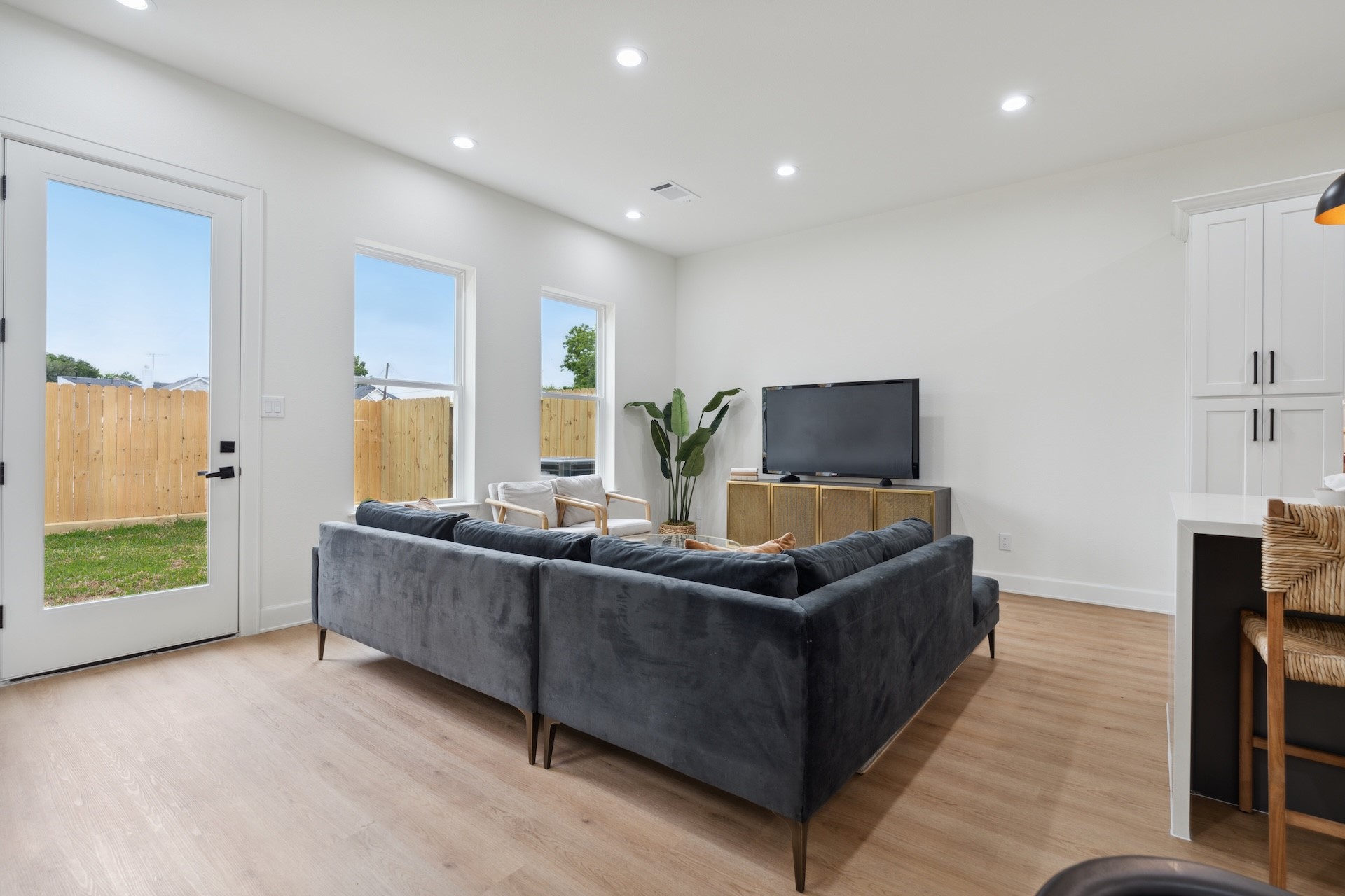 Sunlit living area — glass back door opening to the fenced yard, multiple picture windows, plush sectional arrangement, and light-oak flooring.