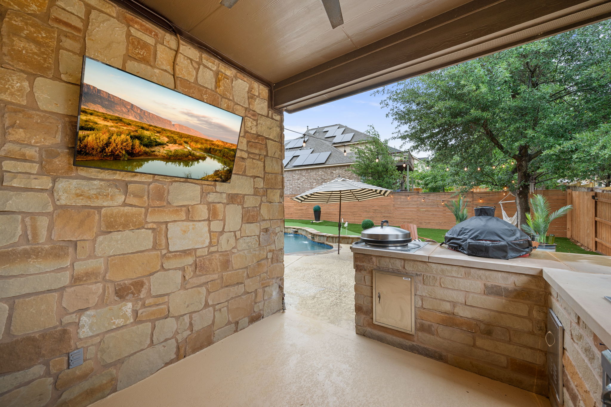 12109 Montclair Bend Austin, TX 78732 - Photo 32 of 39 Covered outdoor patio featuring a stone accent wall with mounted television, a built-in outdoor kitchen with stone base, a ceiling fan, and access to a fenced backyard with a pool