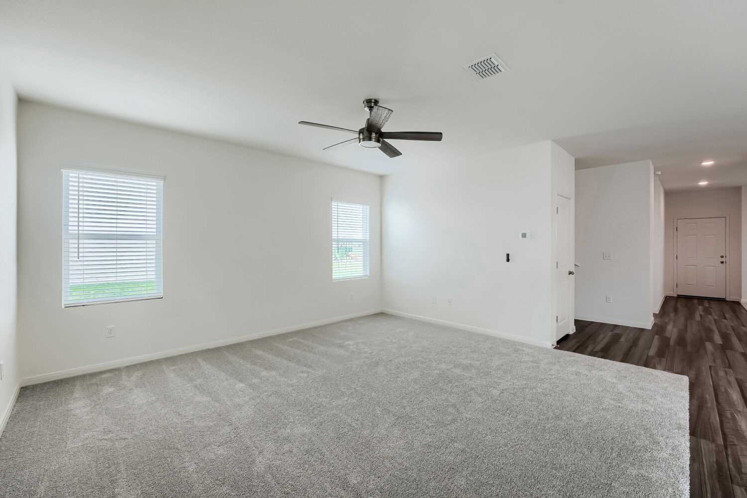 124 Mullen Way Taylor, TX 76574 - Photo 9 of 22 Spacious room featuring light gray carpeting, two windows with white horizontal blinds, a ceiling fan with dark blades, and a transition to dark wood-finish flooring