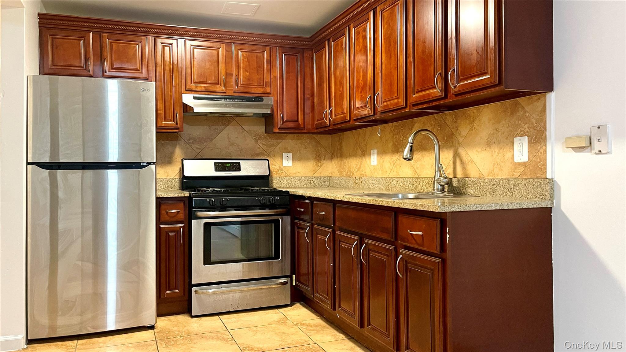 Kitchen with stainless steel appliances, light stone countertops, decorative backsplash, under cabinet range hood, and light tile patterned floors