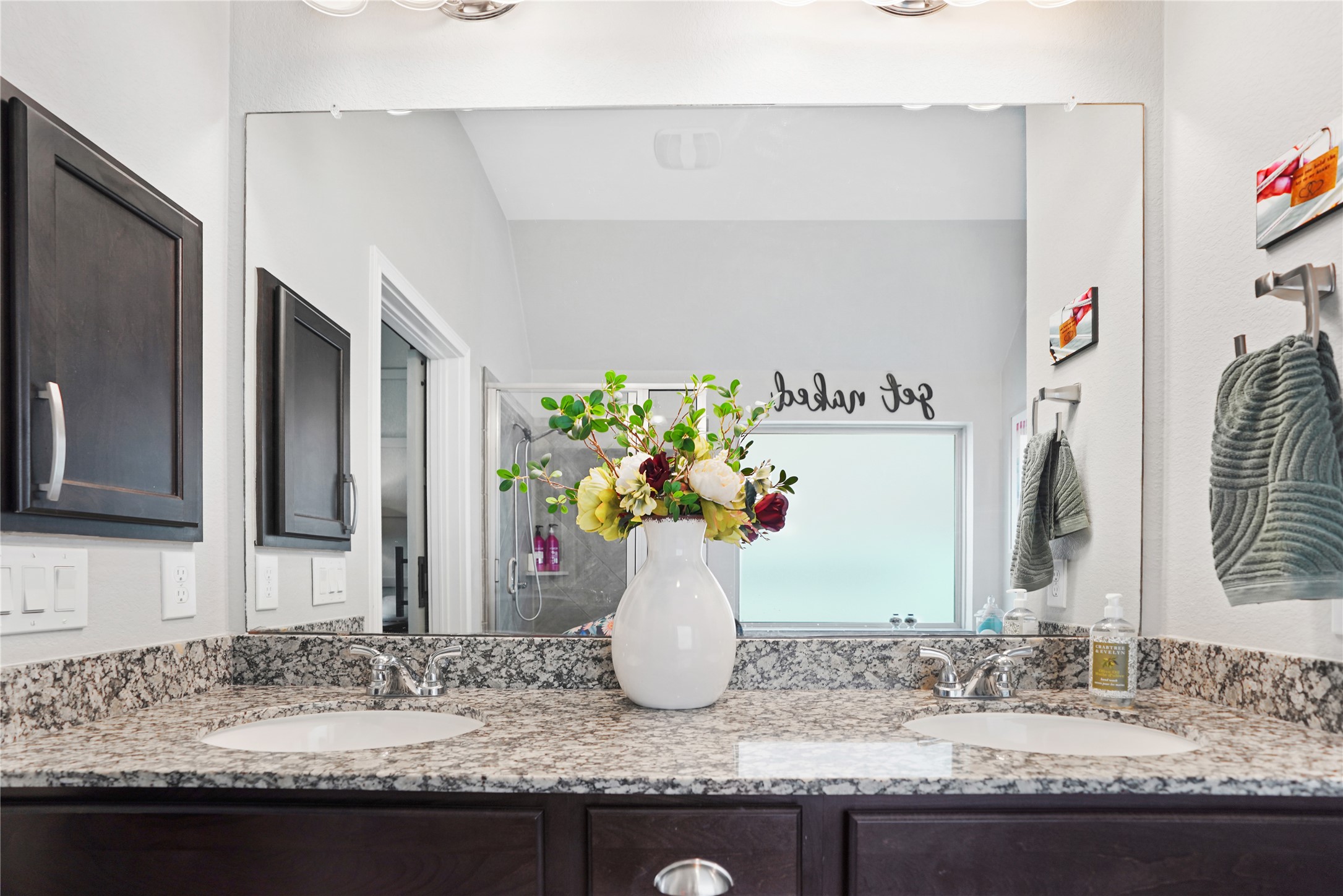 2345 Redwood Ridge Trail Manvel, TX 77578 - Photo 28 of 39 This bathroom features a dual-sink vanity with granite countertops and dark wood cabinets. It includes a large mirror and modern fixtures. A vase with flowers adds a touch of elegance, and there's a walk-in shower visible in the background.