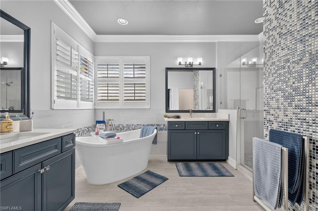 9970 Rookery Circle Estero, FL 33928 - Photo 34 of 50 Freestanding bathtub, dual vanities with dark cabinetry, glass-enclosed shower with mosaic tile, and plantation shutters