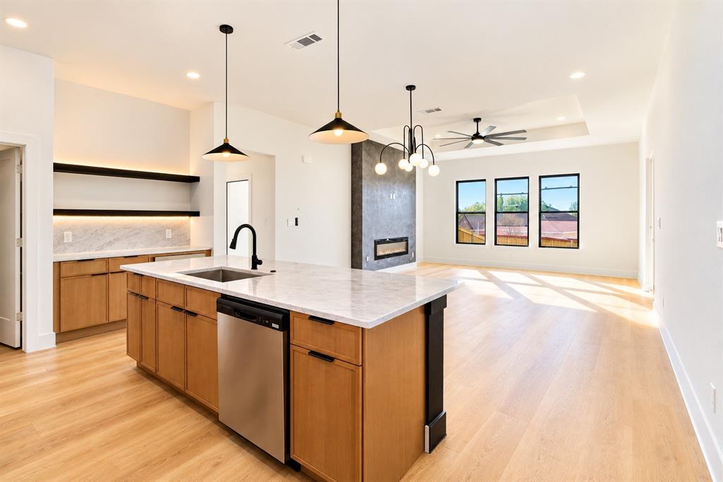 1640 West Wintergreen Road Lancaster, TX 75134 - Photo 5 of 37 Open-concept interior featuring light wood-finish flooring, a kitchen island with a built-in sink and stainless steel dishwasher, a built-in fireplace with a dark surround, and multiple windows
