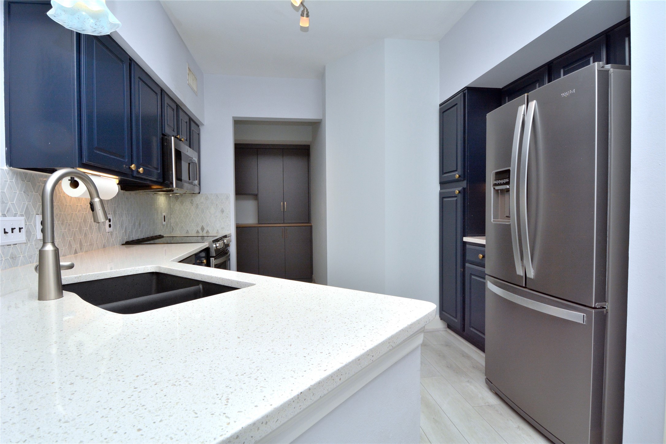 7701 Rialto Boulevard, Unit 124 Austin, TX 78735 - Photo 6 of 22 Kitchen featuring white speckled countertops, a dark undermount sink with a brushed nickel faucet, navy blue cabinetry with gold-toned hardware, stainless steel appliances, and a mosaic tile backsplash