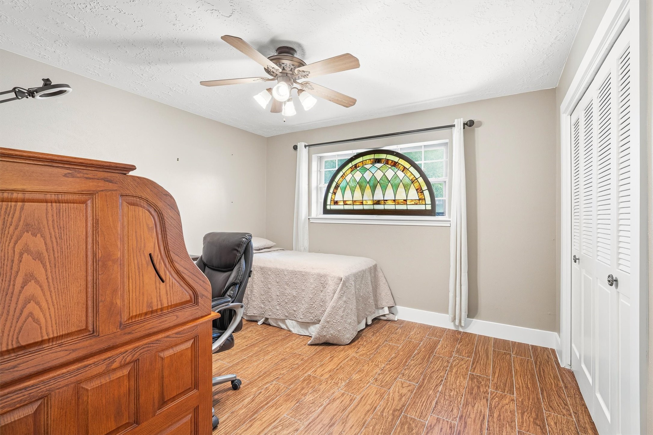 826 North Rivershire Drive Conroe, TX 77304 - Photo 25 of 46 Bedroom 3 has a detail you won't find in most homes at any price point — a stained glass arched window that fills the room with color and light in a way that feels genuinely special. Wood look flooring, ceiling fan, and a louvered closet complete the space.