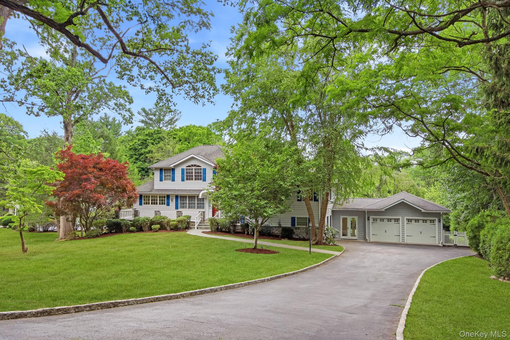 26 Century Ridge Road Purchase, NY 10577 - Photo 2 of 50 Expansive front yard featuring a light gray facade with blue window shutters, a prominent two-car garage, mature trees, and a paved driveway