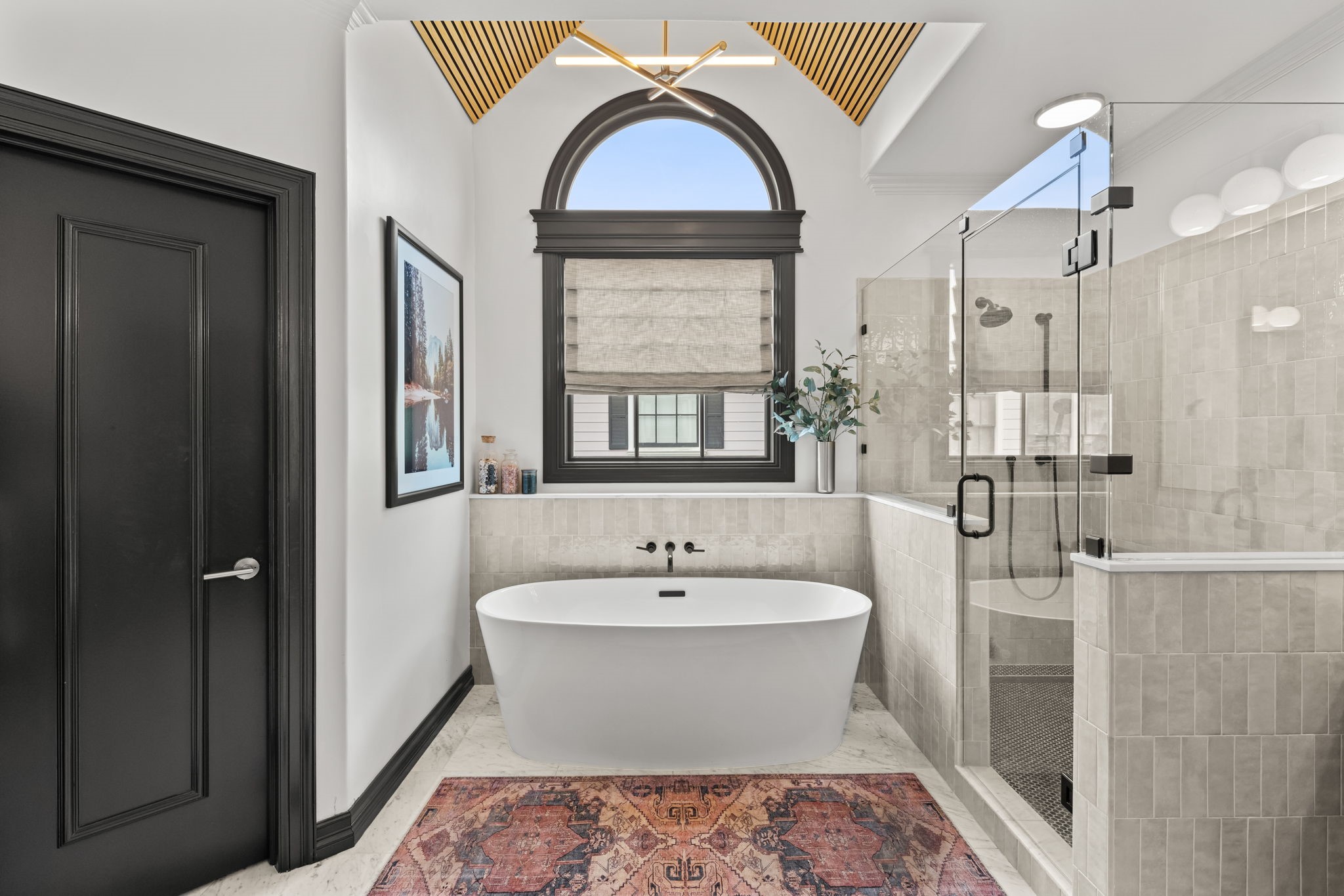 520 Oxford Street, Unit F Houston, TX 77007 - Photo 20 of 38 A dramatic arched window frames the freestanding soaking tub, which is positioned beneath a vaulted ceiling feature with a wood slat inlay and a linear chandelier creating an architectural focal point that elevates the entire space. The adjacent walk-in glass shower features floor-to-ceiling handmade tile and matte black fixtures.