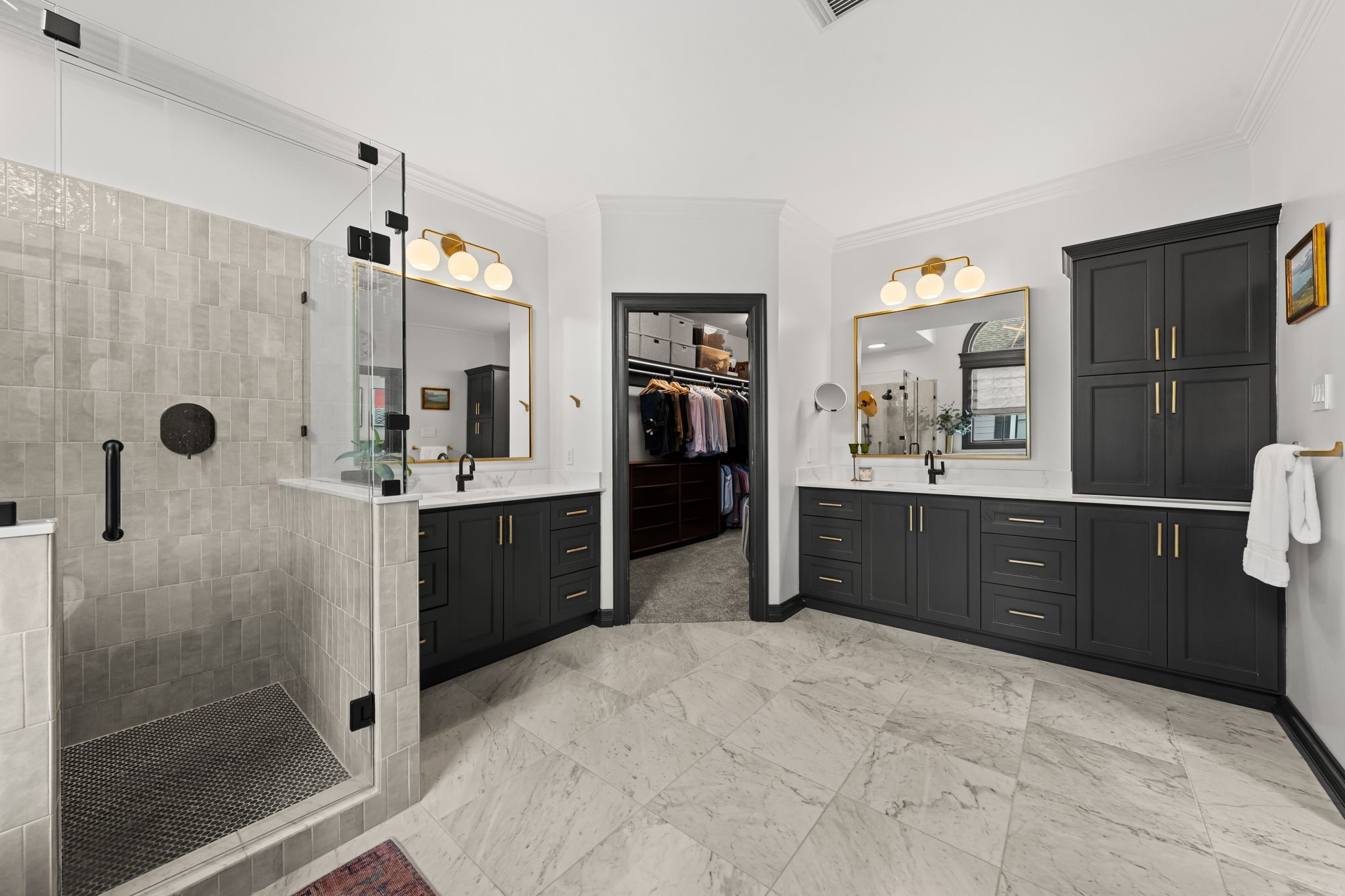 520 Oxford Street, Unit F Houston, TX 77007 - Photo 22 of 38 Dual dark vanities with marble-look countertops, gold-framed mirrors, and globe sconce lighting line both walls of this luxurious primary bath, offering generous storage and counter space. The open doorway between them leads directly into the custom walk-in closet, creating a seamless dressing suite experience.