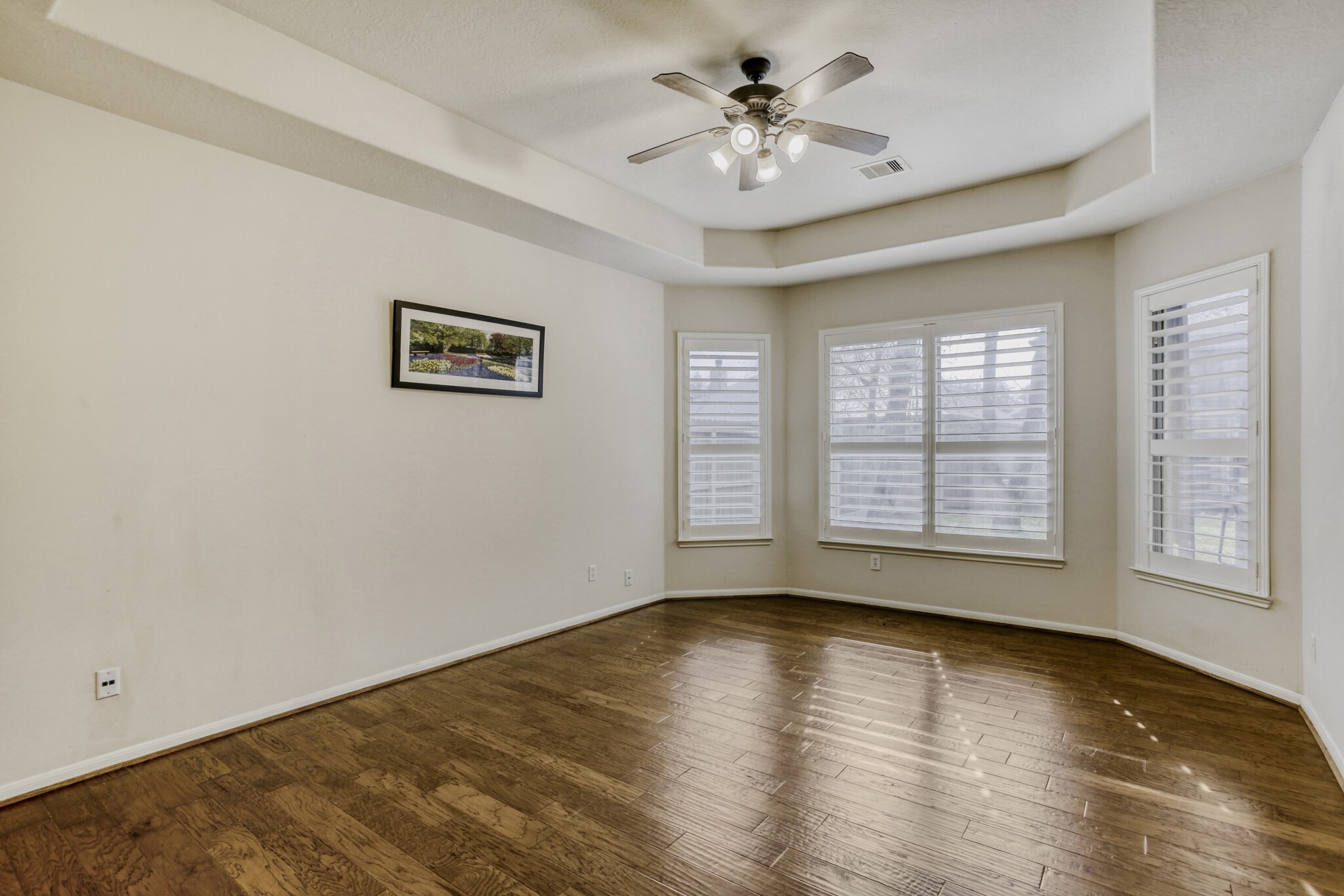 19303 Valiant Woods Drive Spring, TX 77379 - Photo 15 of 27 Discover the master bedroom's serene elegance, featuring rich wood flooring, abundant natural light through stylish shutters, and a chic tray ceiling with a modern fan for ultimate comfort.