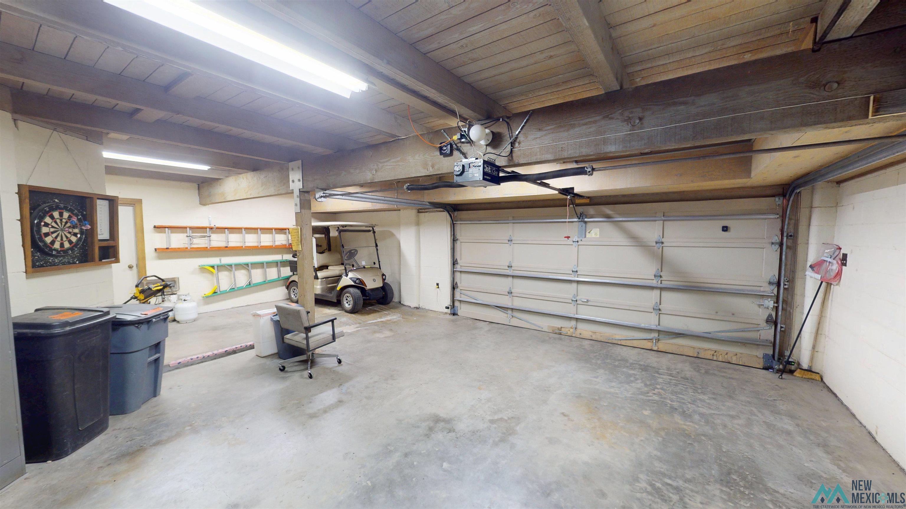 39 Alta Vista Rociada, NM 87742 - Photo 36 of 54 Spacious garage featuring a concrete floor, exposed wood beam ceiling, multiple fluorescent light fixtures, a large overhead garage door with opener, and built-in wall shelving