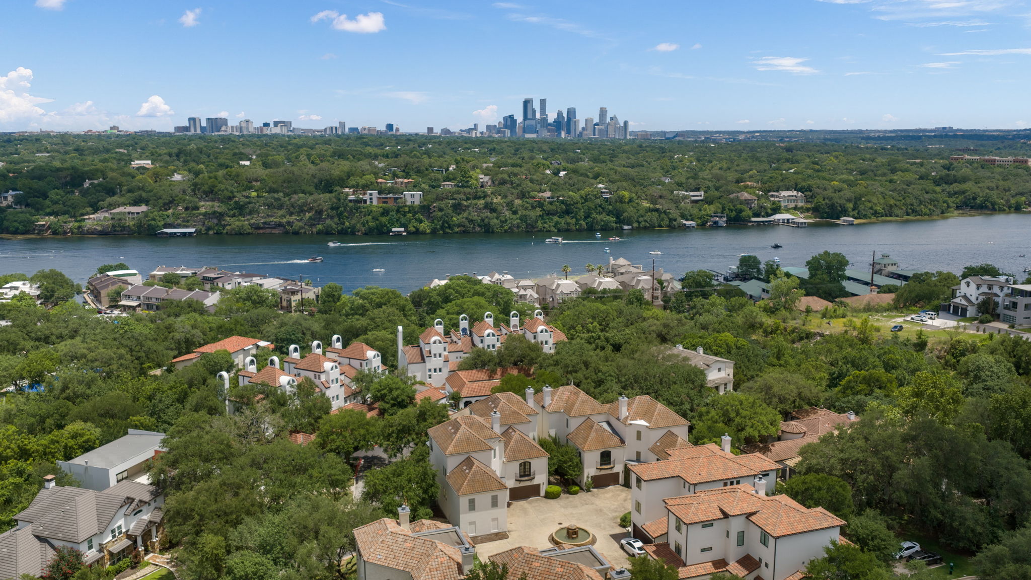 1910 Rue De St Tropez, Unit 6 Austin, TX 78746 - Photo 2 of 37 An aerial perspective reveals a serene waterfront community nestled amidst lush greenery with a cityscape horizon.
