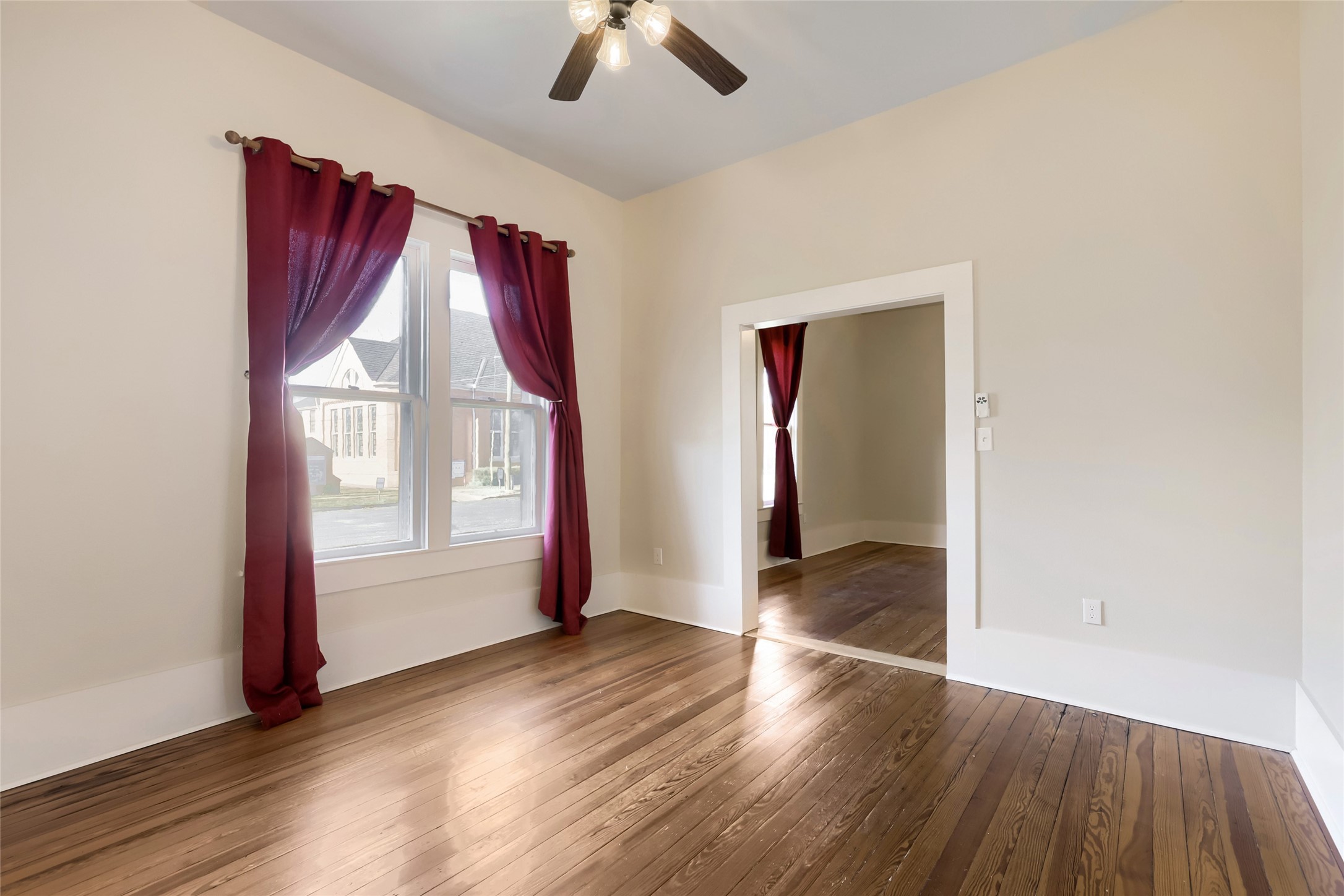 208 North Ave B Elgin, TX 78621 - Photo 5 of 29 Bright room with large windows, red curtains, and hardwood floors. Features a ceiling fan and an open doorway connecting to another space, offering a warm and inviting atmosphere.