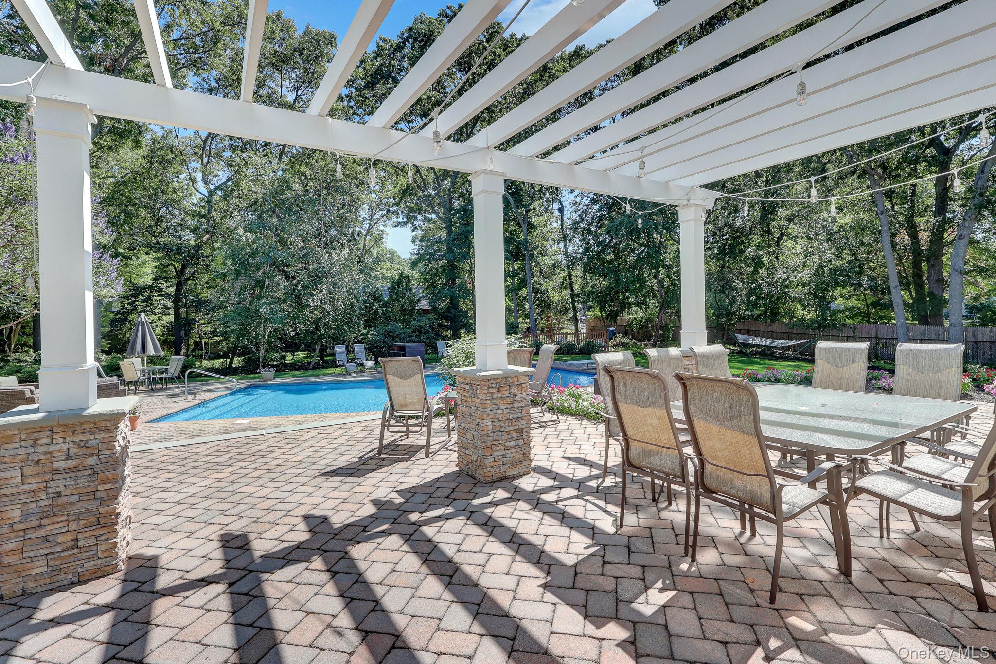 24 Fox Road East Setauket, NY 11733 - Photo 39 of 48 Expansive paver patio with a white pergola featuring string lighting and stone-clad pillars