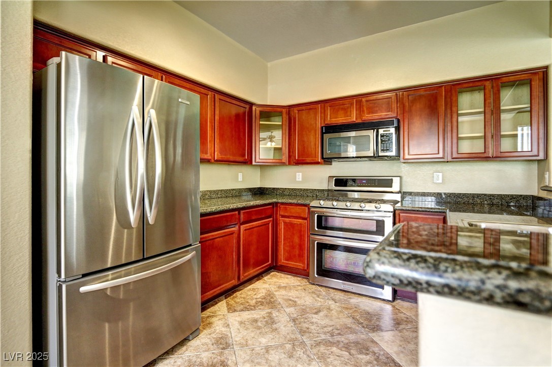10225 King Henry Avenue, Unit 104 Las Vegas, NV 89144 - Photo 8 of 24 Kitchen featuring reddish brown cabinets, appliances with stainless steel finishes, glass insert cabinets, dark stone counters, and light tile patterned floors