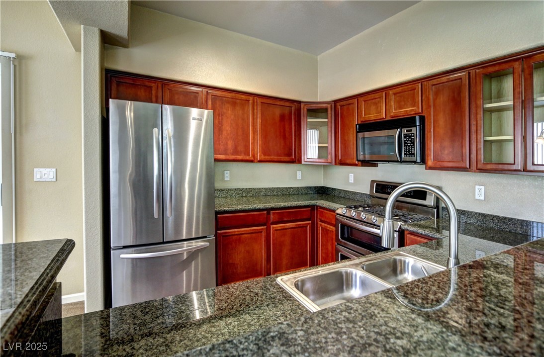 10225 King Henry Avenue, Unit 104 Las Vegas, NV 89144 - Photo 10 of 24 Kitchen featuring appliances with stainless steel finishes, glass insert cabinets, a textured wall, reddish brown cabinets, and dark stone countertops